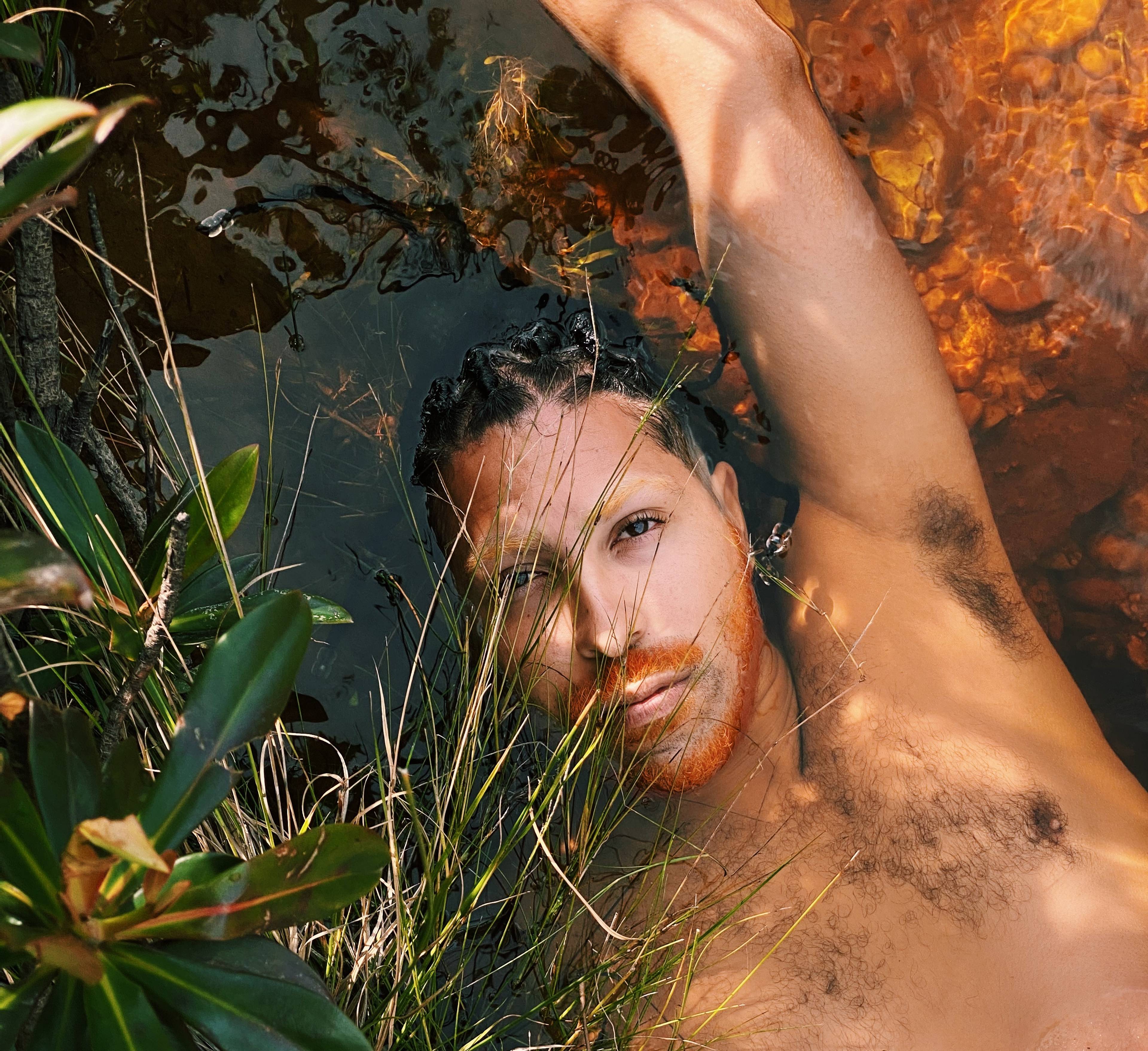A person with braided hair and facial hair lies partially submerged in water, surrounded by green plants. Sunlight creates dappled shadows on their skin.