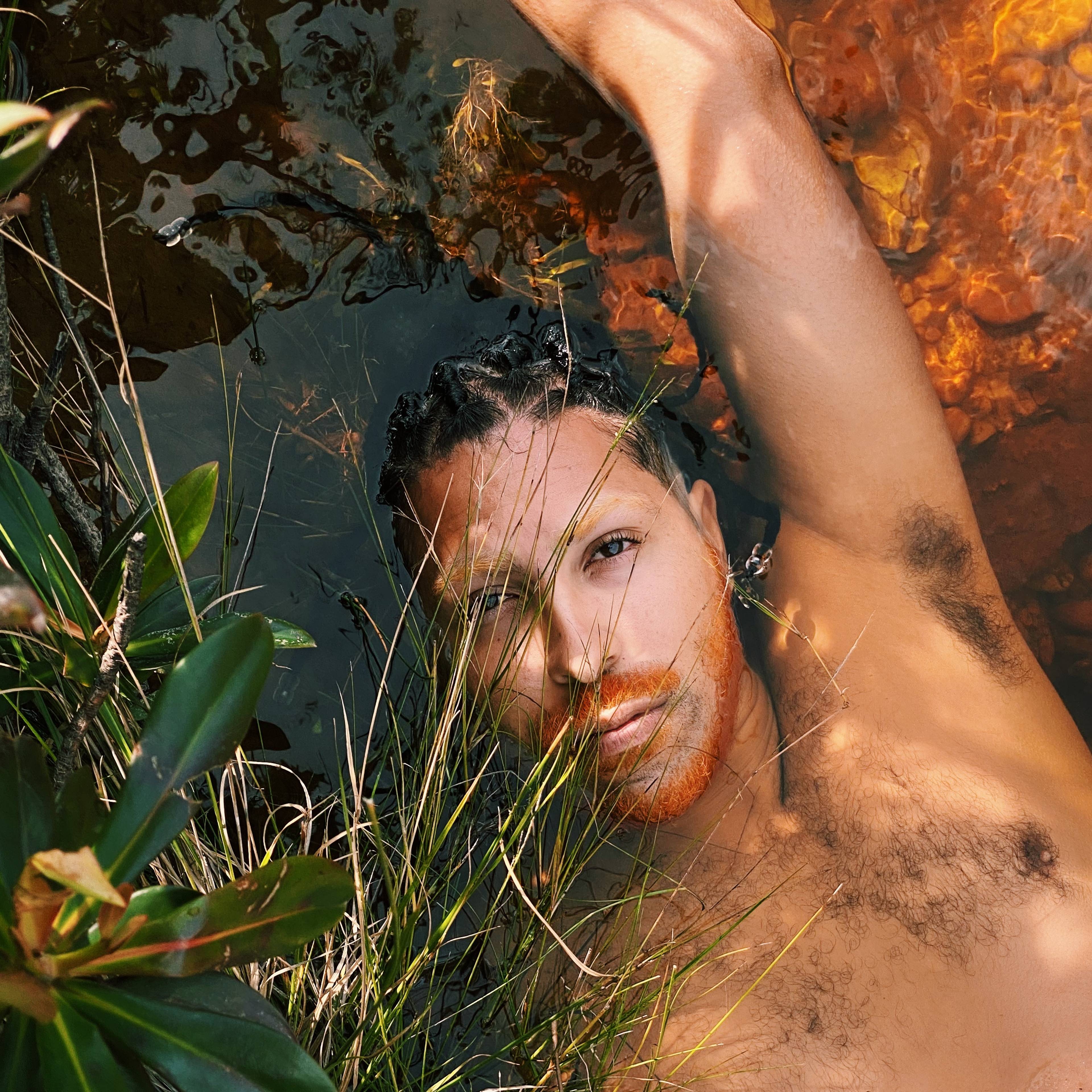 A person with braided hair and facial hair lies partially submerged in water, surrounded by green plants. Sunlight creates dappled shadows on their skin.