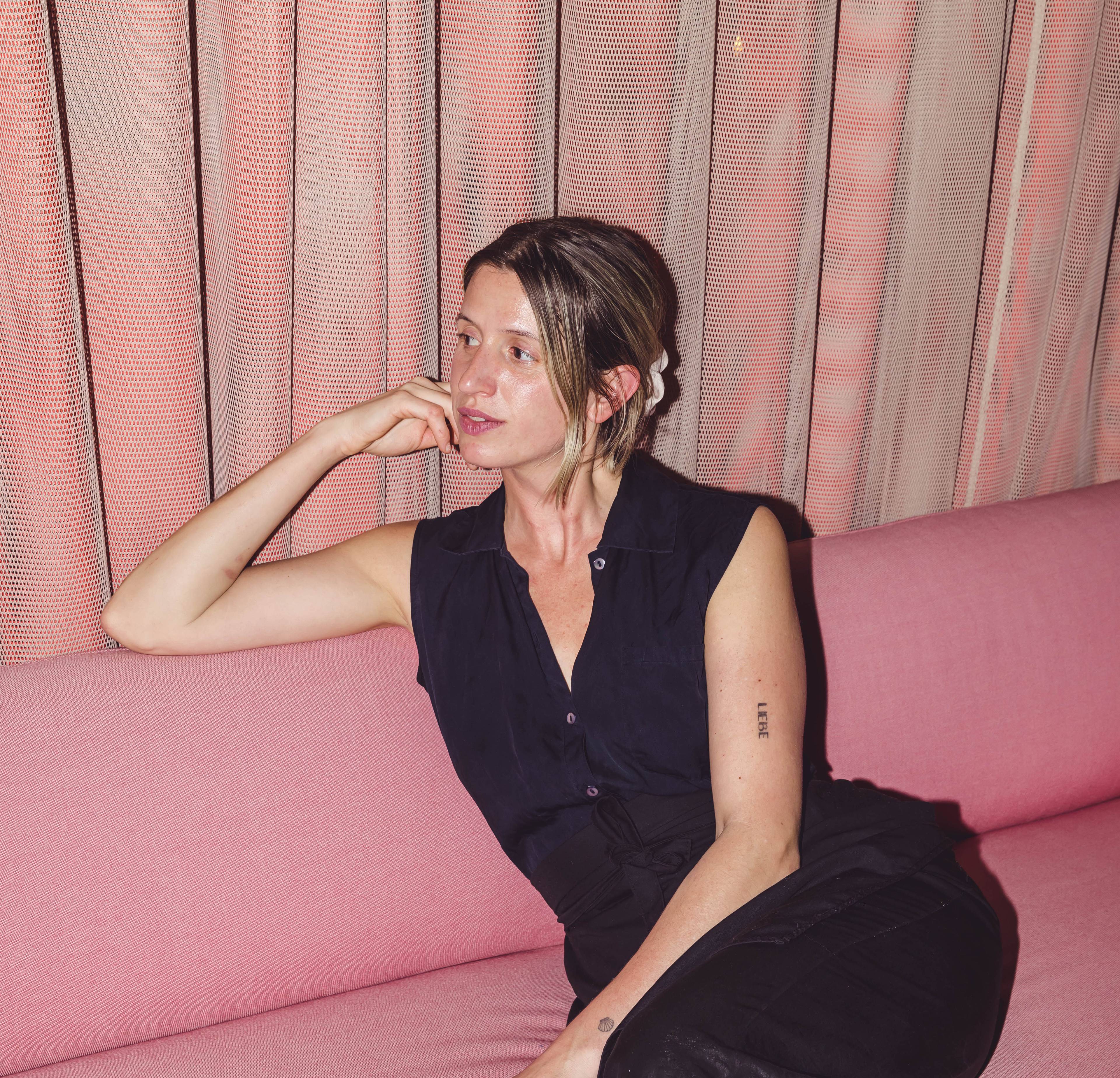 A person with tied-up hair and a sleeveless black top sits on a pink sofa, their head resting on their hand. Pink curtains are in the background, creating a soft ambiance. The person has a calm and relaxed expression.