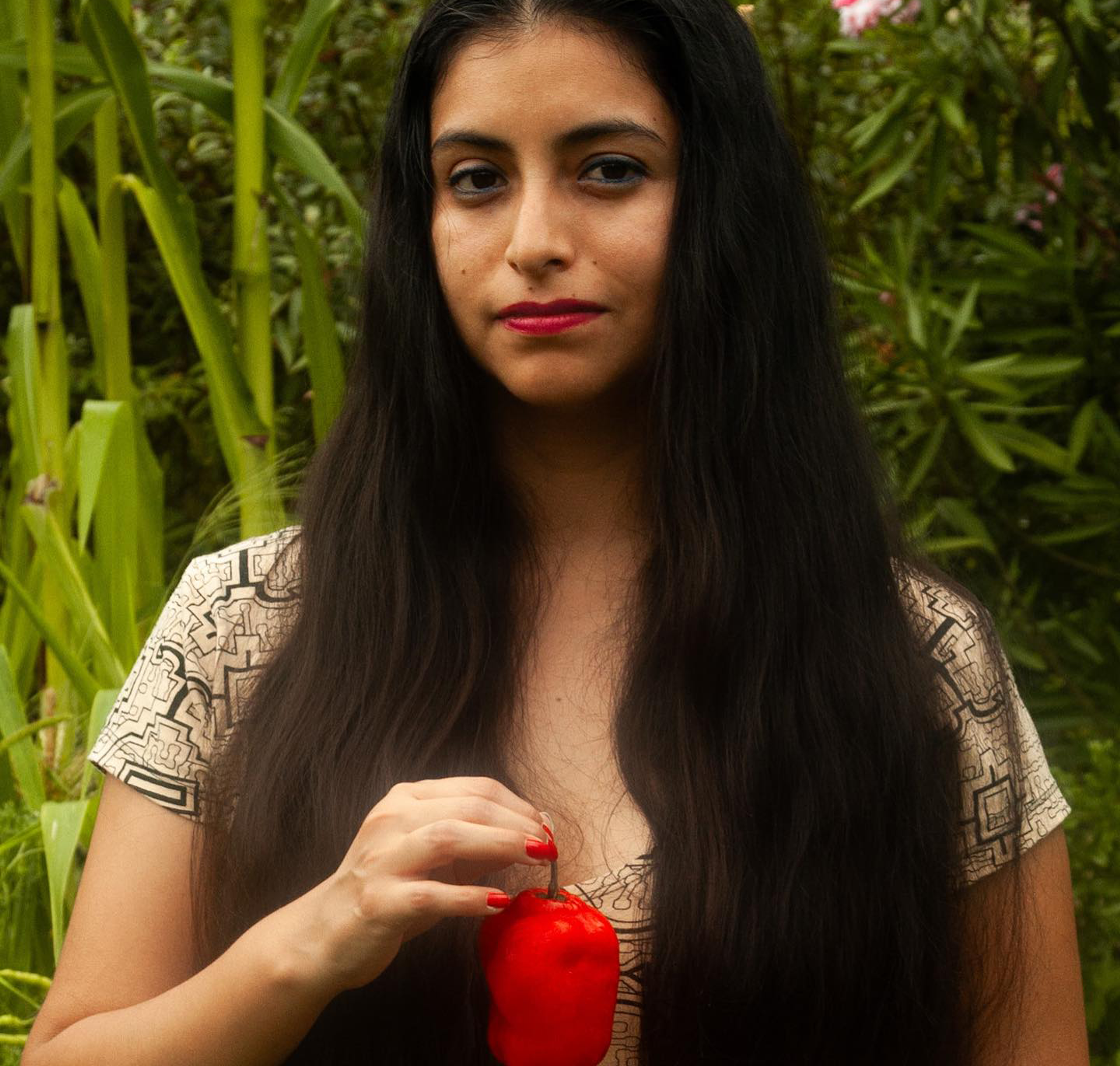 Eine Person mit langen dunklen Haaren und rotem Lippenstift hält eine kleine rote Paprika. Sie trägt ein gemustertes Oberteil und steht im Freien mit üppig grünem Laub im Hintergrund.