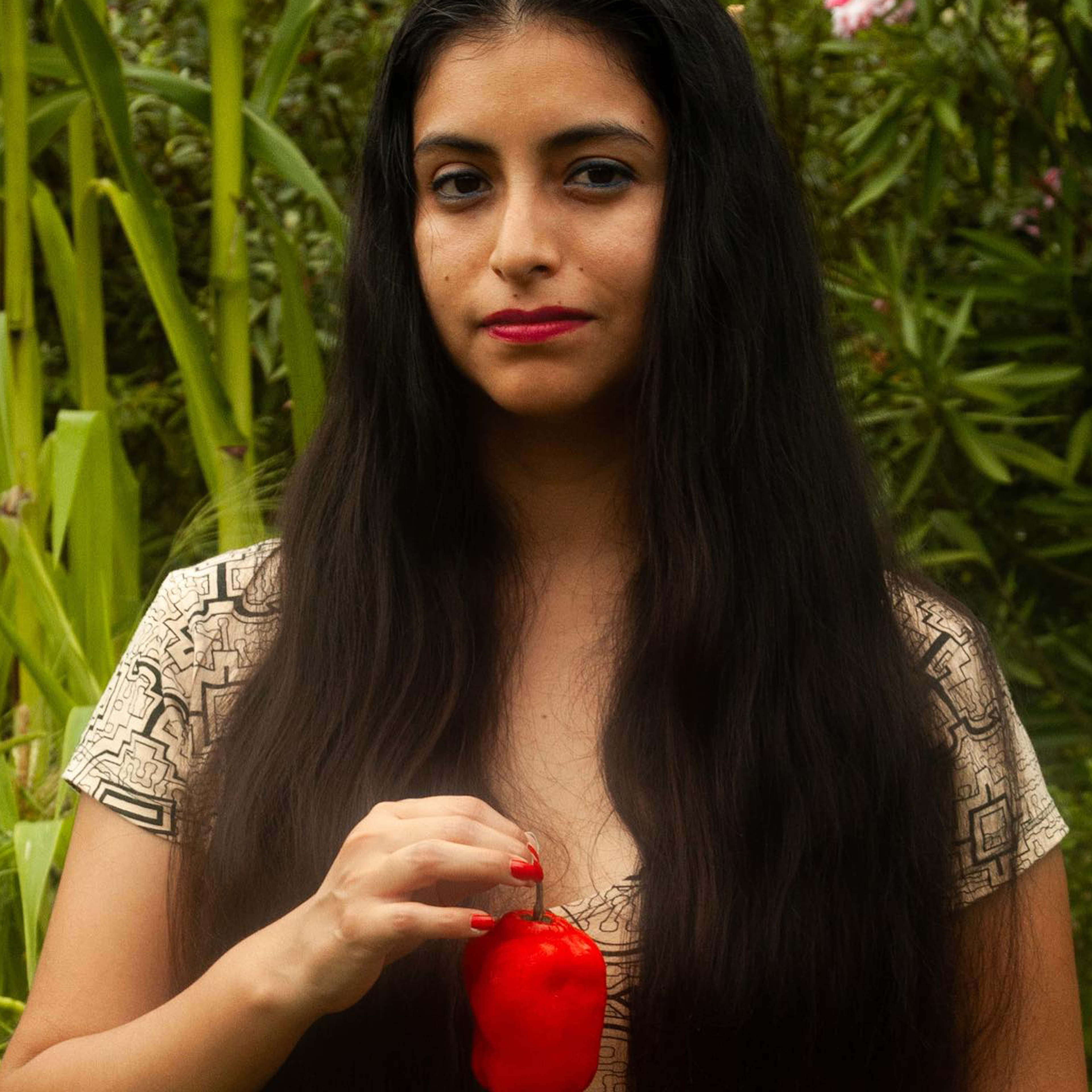 A person with long dark hair and red lipstick holds a small red bell pepper. They are wearing a patterned top and standing outdoors with lush green foliage in the background.