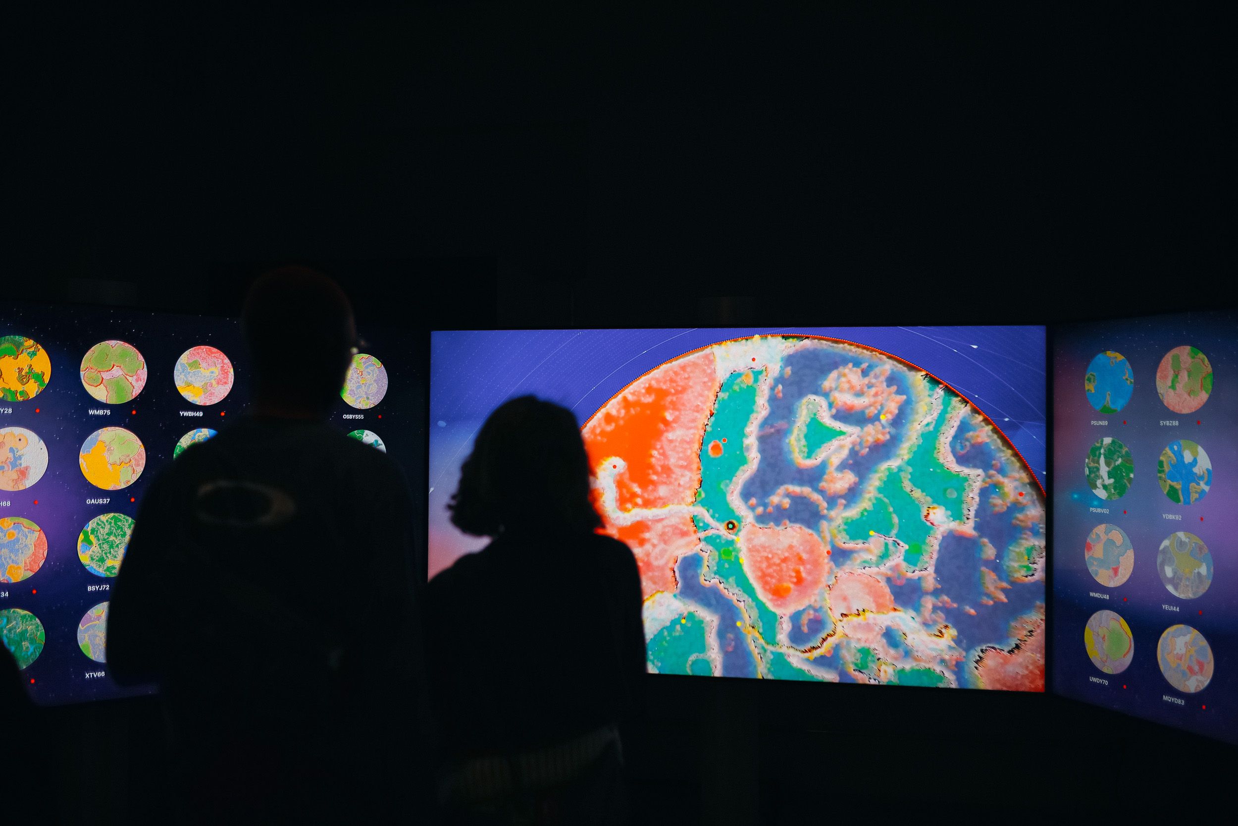 Zwei Personen stehen als Silhouetten vor drei großen Bildschirmen, auf denen farbenfrohe digitale Welten zu sehen sind. Der mittlere Bildschirm zeigt eine Nahaufnahme einer lebhaften abstrakten Landschaft in Blau, Grün, Pink und Orange, während auf den seitlichen Bildschirmen mehrere kreisförmige Weltansichten dargestellt sind.