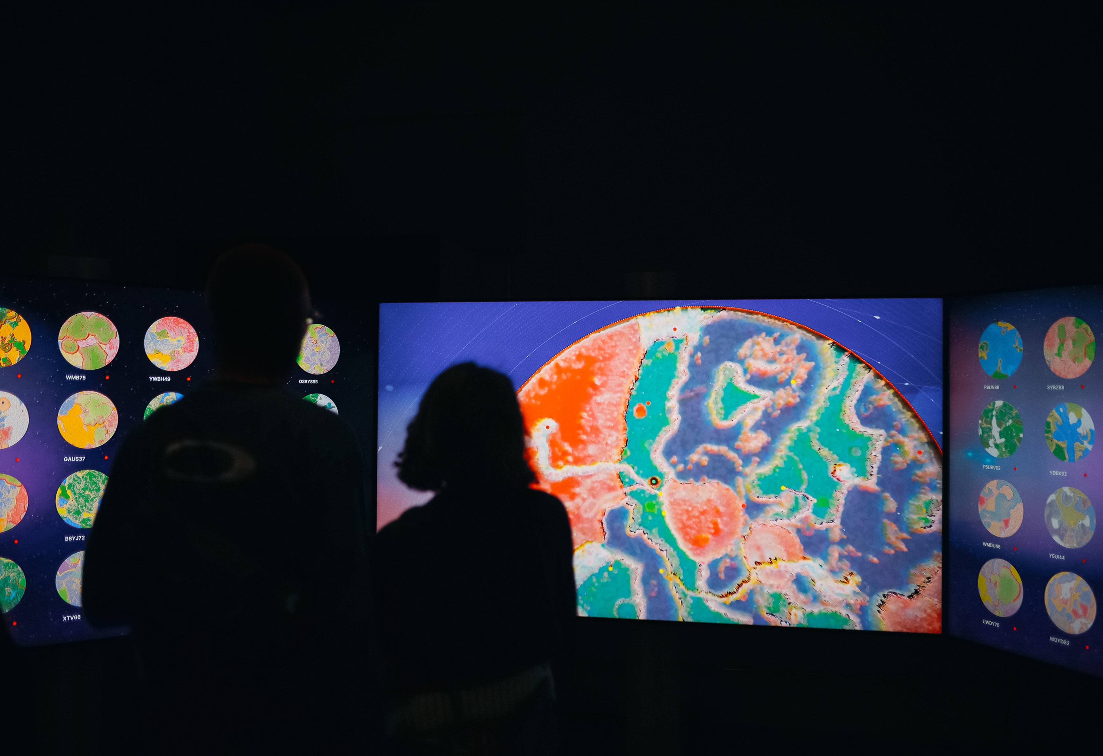 Zwei Personen stehen als Silhouetten vor drei großen Bildschirmen, auf denen farbenfrohe digitale Welten zu sehen sind. Der mittlere Bildschirm zeigt eine Nahaufnahme einer lebhaften abstrakten Landschaft in Blau, Grün, Pink und Orange, während auf den seitlichen Bildschirmen mehrere kreisförmige Weltansichten dargestellt sind.