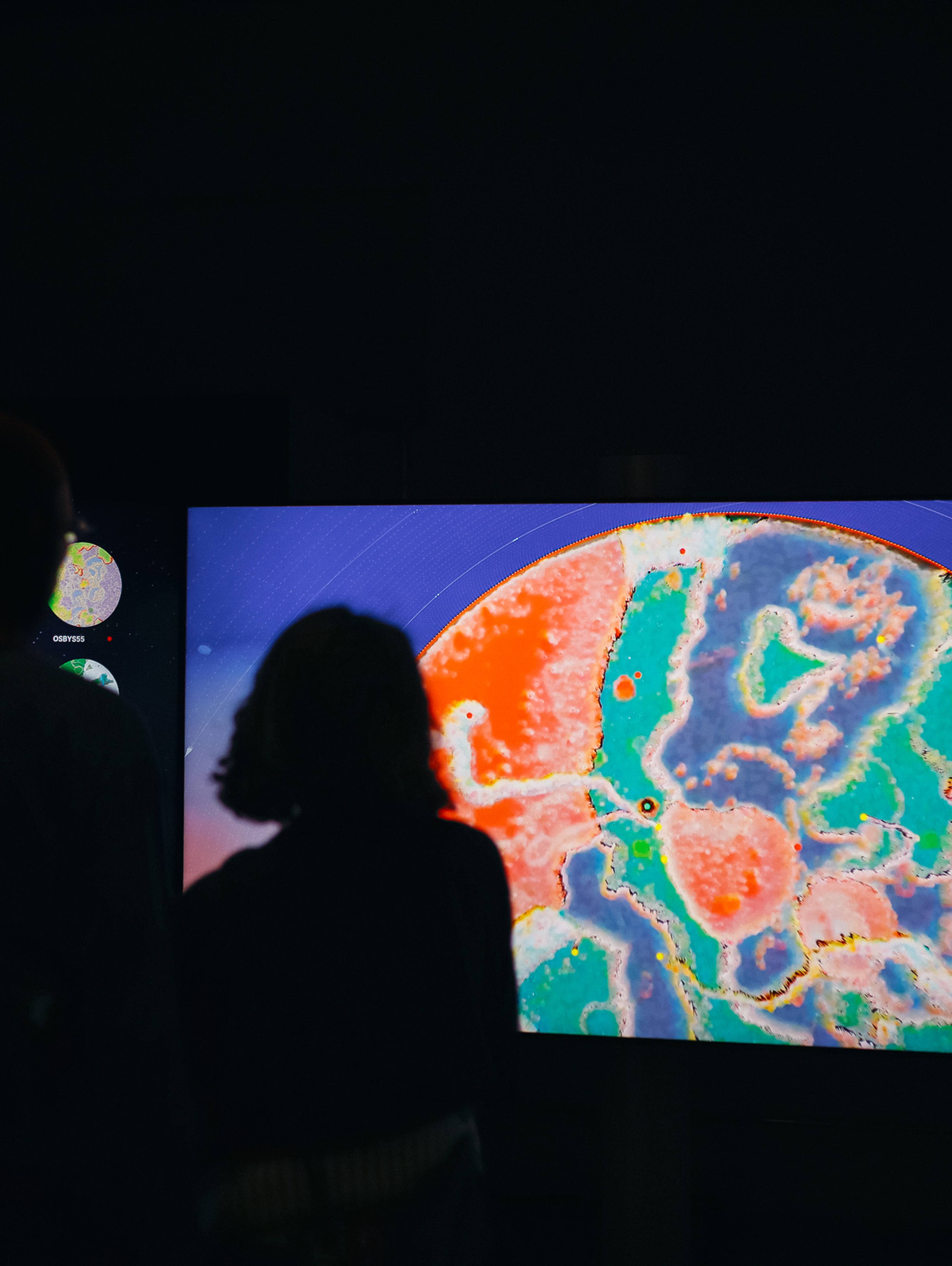 Zwei Personen stehen als Silhouetten vor drei großen Bildschirmen, auf denen farbenfrohe digitale Welten zu sehen sind. Der mittlere Bildschirm zeigt eine Nahaufnahme einer lebhaften abstrakten Landschaft in Blau, Grün, Pink und Orange, während auf den seitlichen Bildschirmen mehrere kreisförmige Weltansichten dargestellt sind.