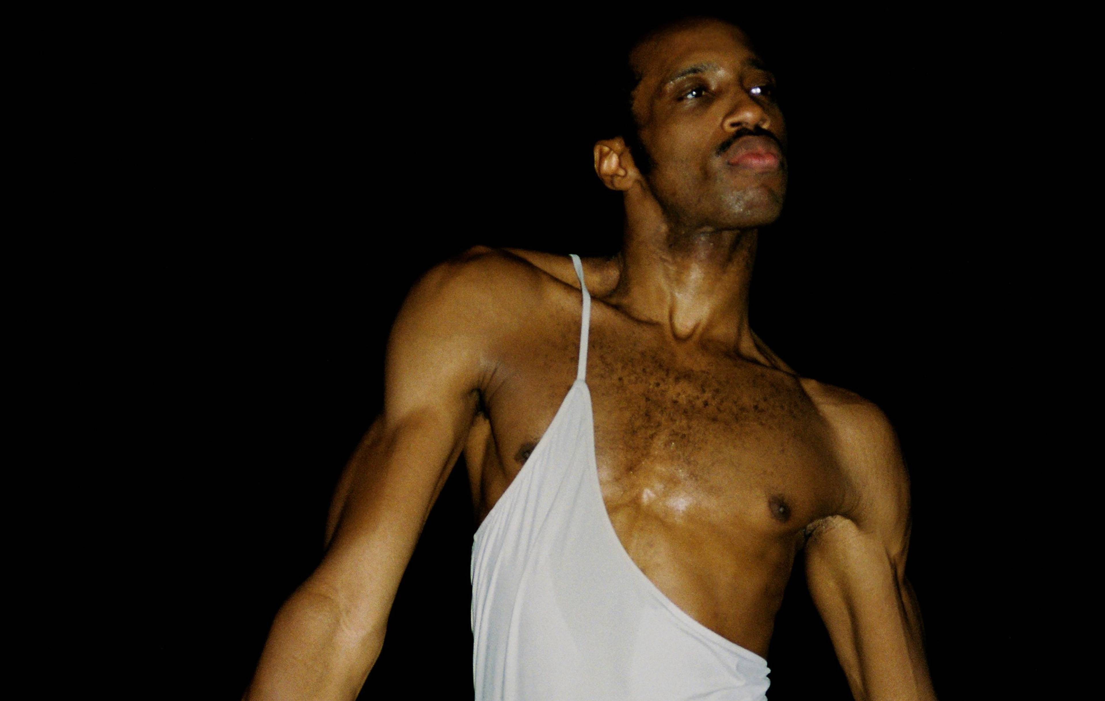 A person wearing a partially off-shoulder white tank top is standing against a dark background. They have a contemplative expression and an athletic build.