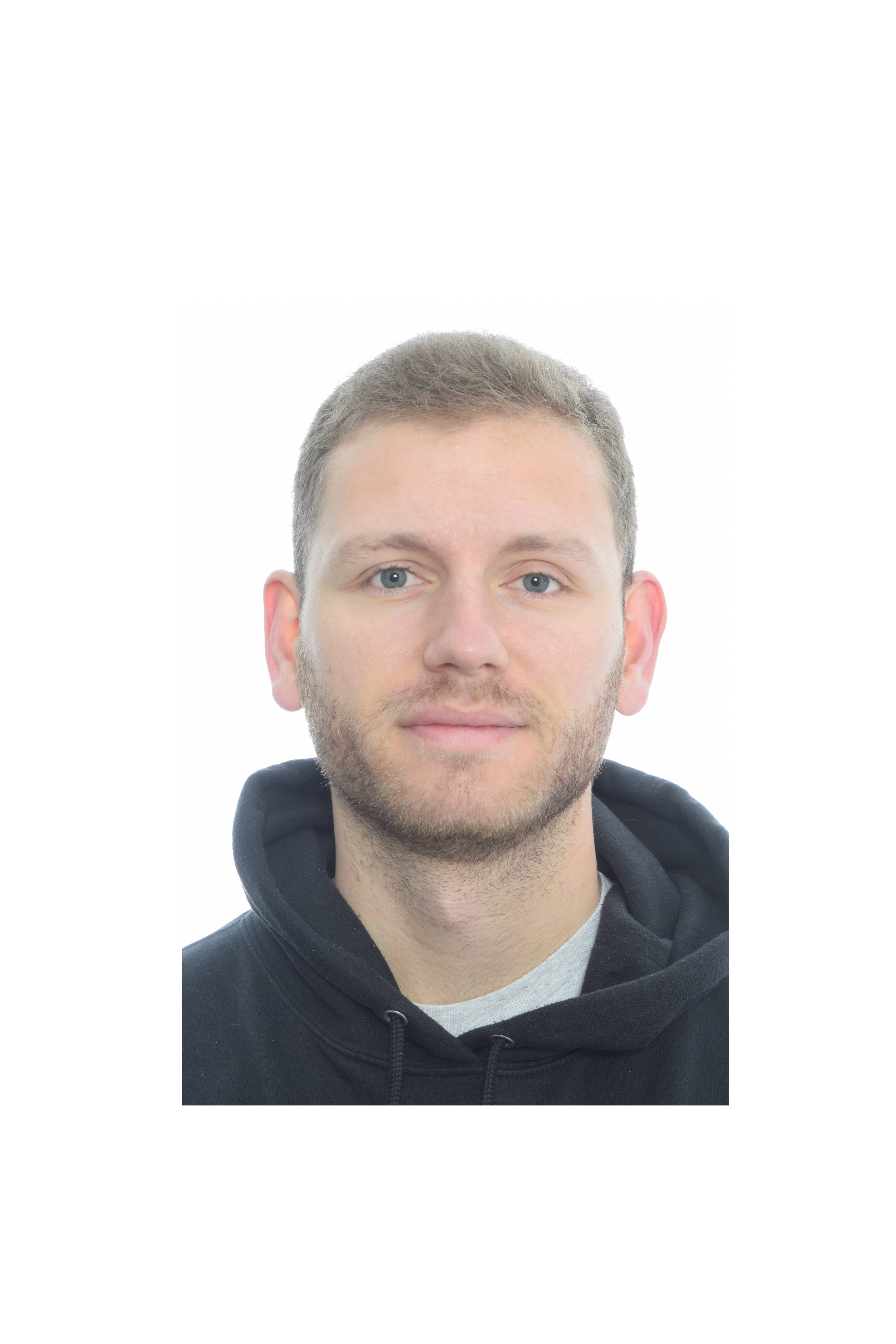 A person with short light brown hair and a trimmed beard looks at the camera with a neutral expression, wearing a dark hoodie against a plain white background.