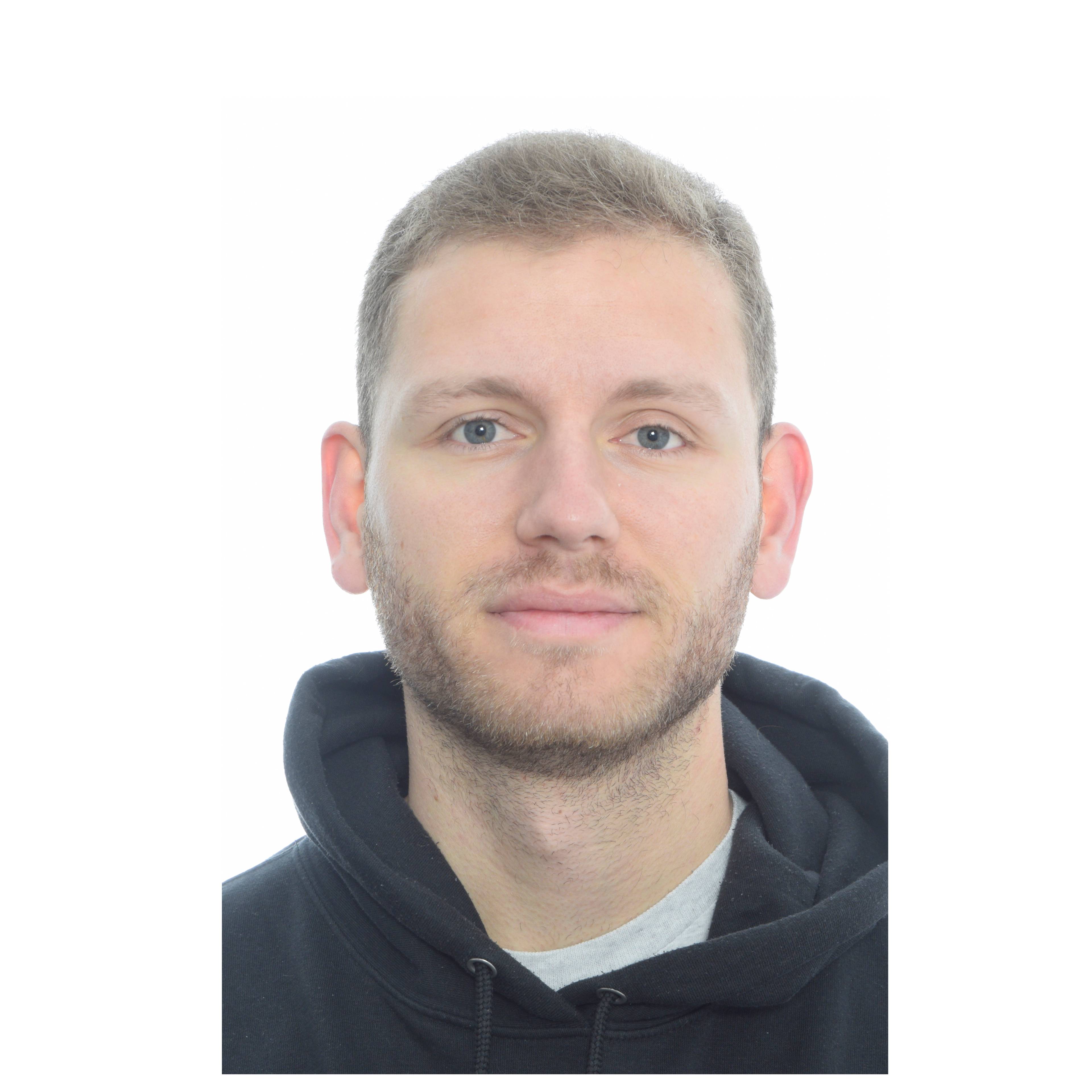 A person with short light brown hair and a trimmed beard looks at the camera with a neutral expression, wearing a dark hoodie against a plain white background.