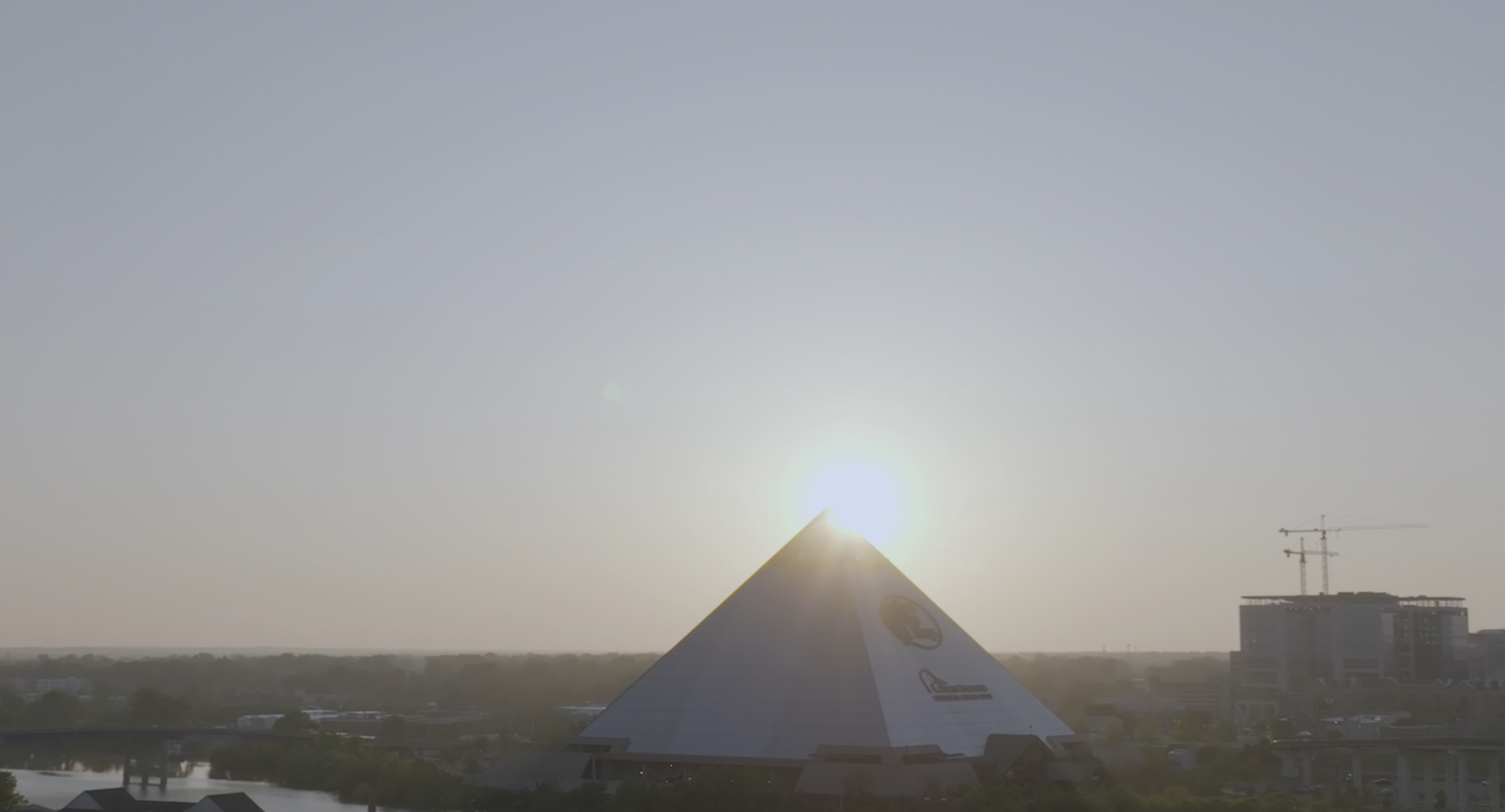Ein großes pyramidenförmiges Glasgebäude steht unter klarem Himmel bei Sonnenaufgang. Die Sonne befindet sich direkt über der Spitze der Pyramide und wirft ein sanftes Licht auf die Szene. Im Hintergrund sind einige Gebäude und ein Kran zu sehen.