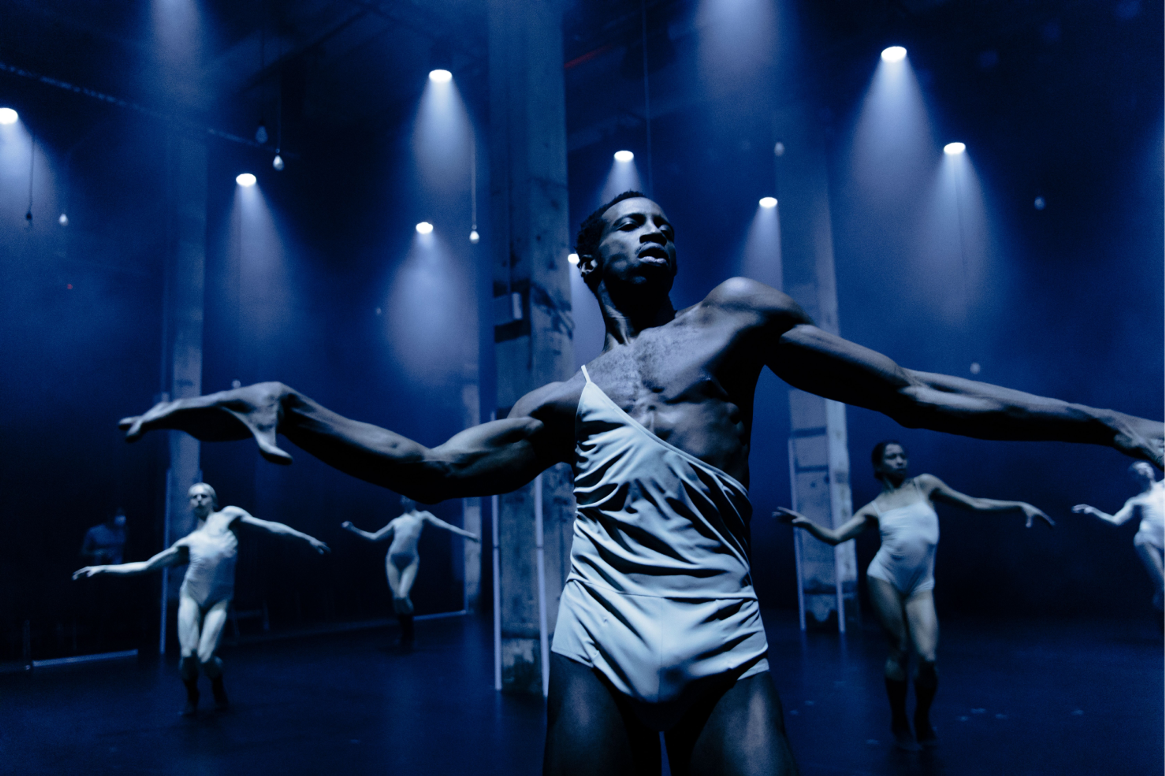 L-E-V dance company dancers engaging in a coordinated movement with arms stretched out under blue-tinged spotlights.