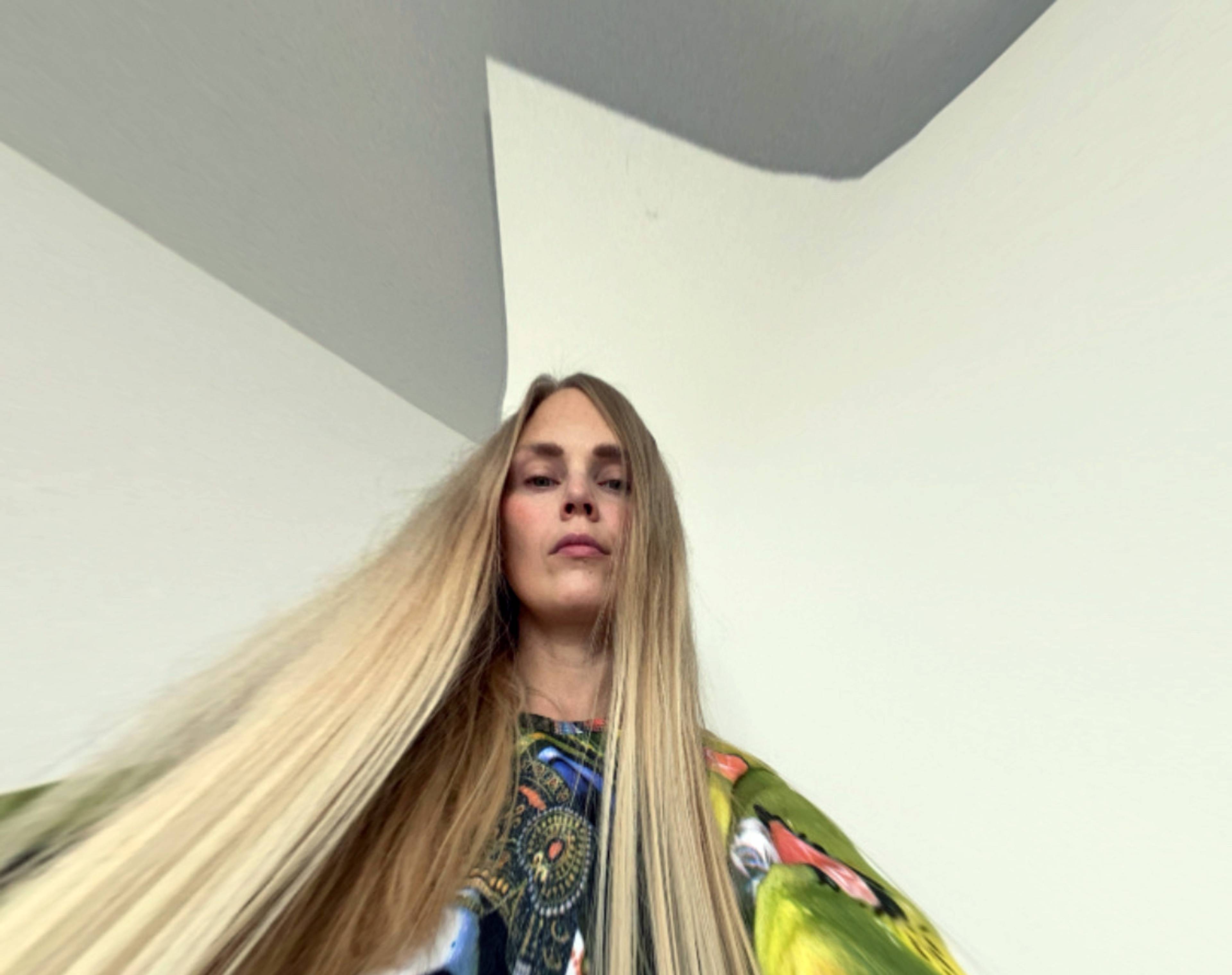 A person with very long, stretched hair stands indoors, wearing a colorful, patterned outfit. The camera angle is from below, creating a distorted perspective.