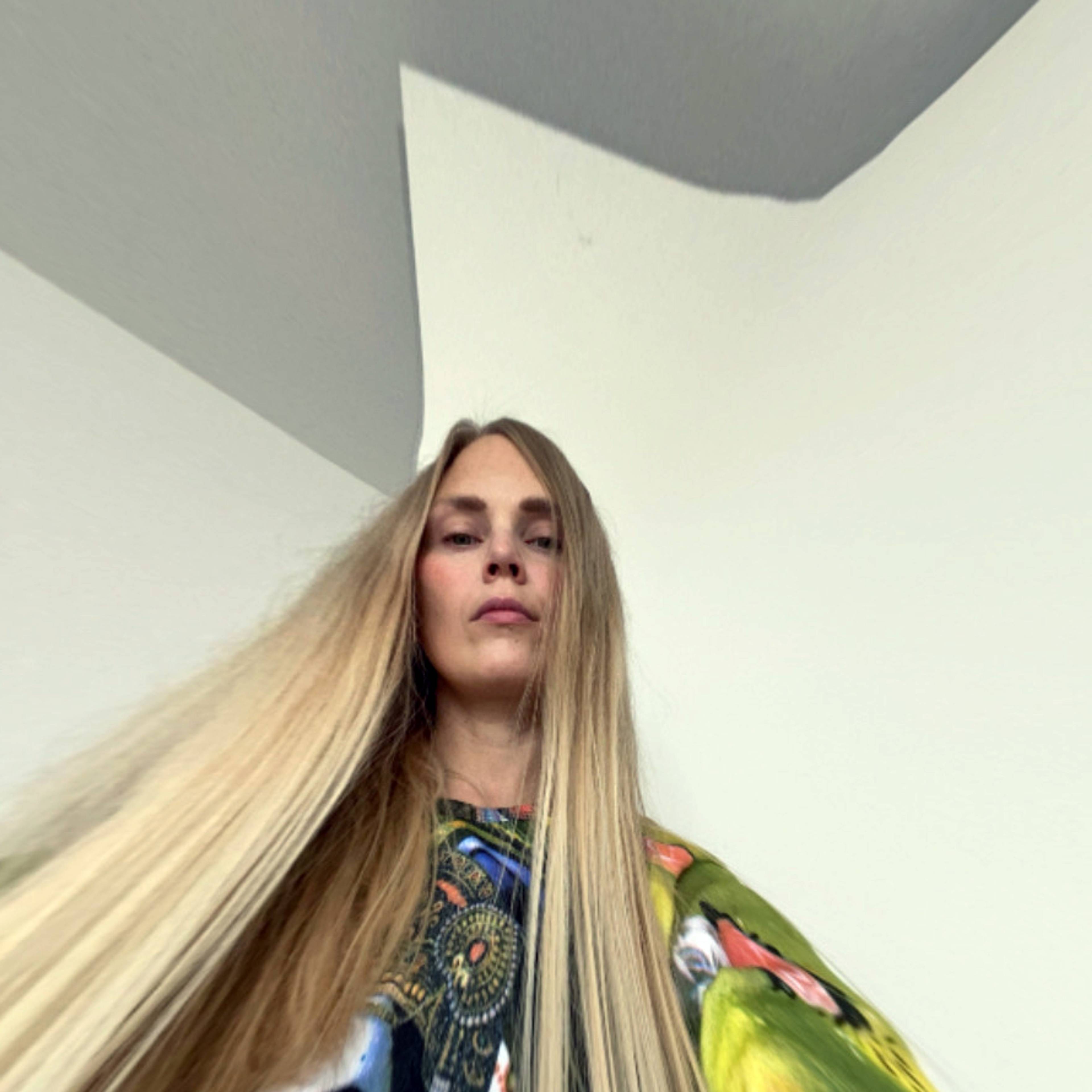Eine Person mit sehr langem, gestrecktem Haar steht in einem Innenraum und trägt ein bunt gemustertes Outfit. Der Kamerawinkel ist von unten, wodurch eine verzerrte Perspektive entsteht.