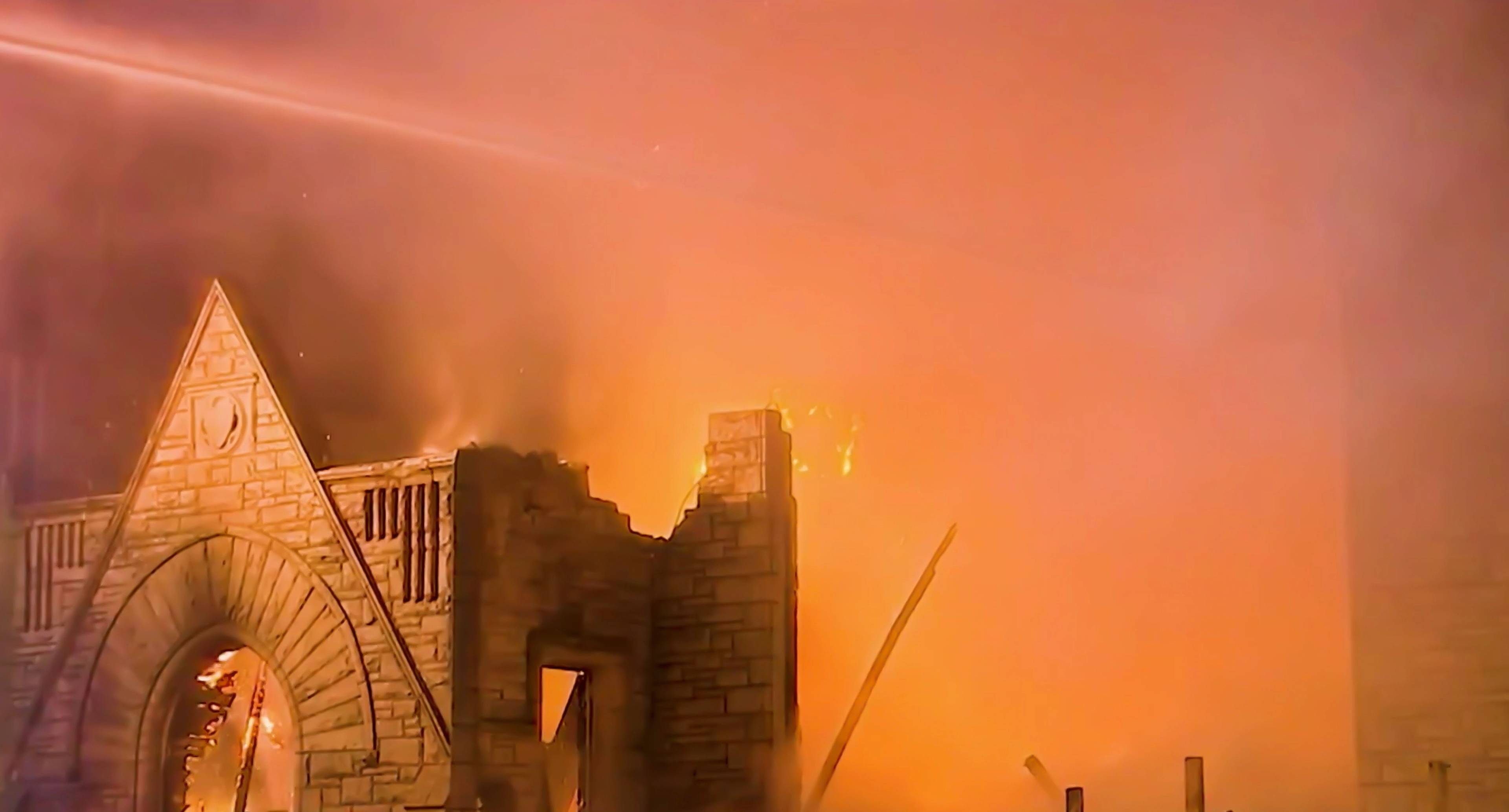 Ein kirchenähnliches Gebäude aus Stein steht in Flammen; dichter orangener Rauch und Feuer steigen nachts aus der Struktur auf.