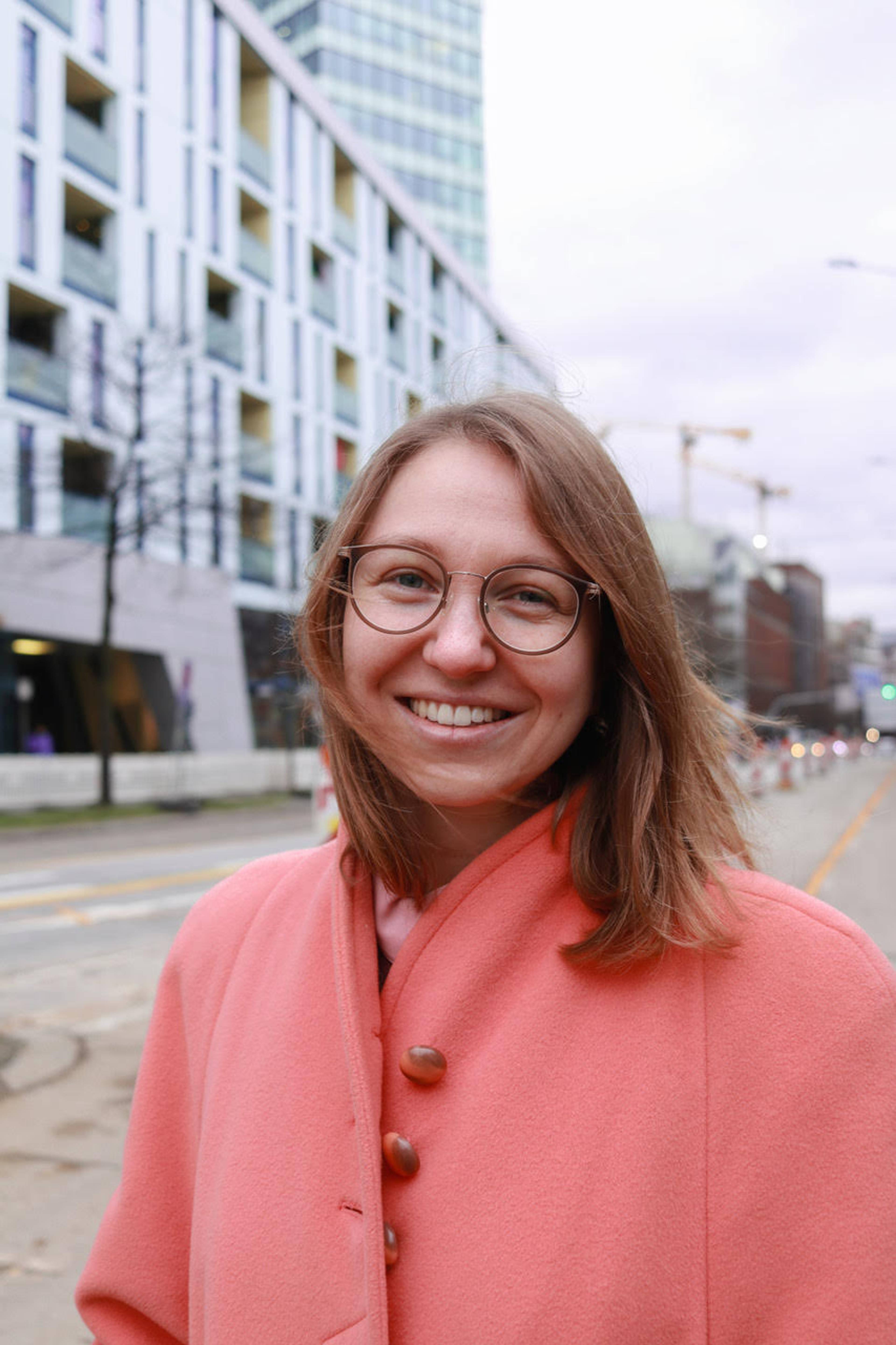 A person wearing glasses and a coral coat smiles at the camera. They stand on a city street with modern buildings and construction cranes in the background. Its an overcast day.