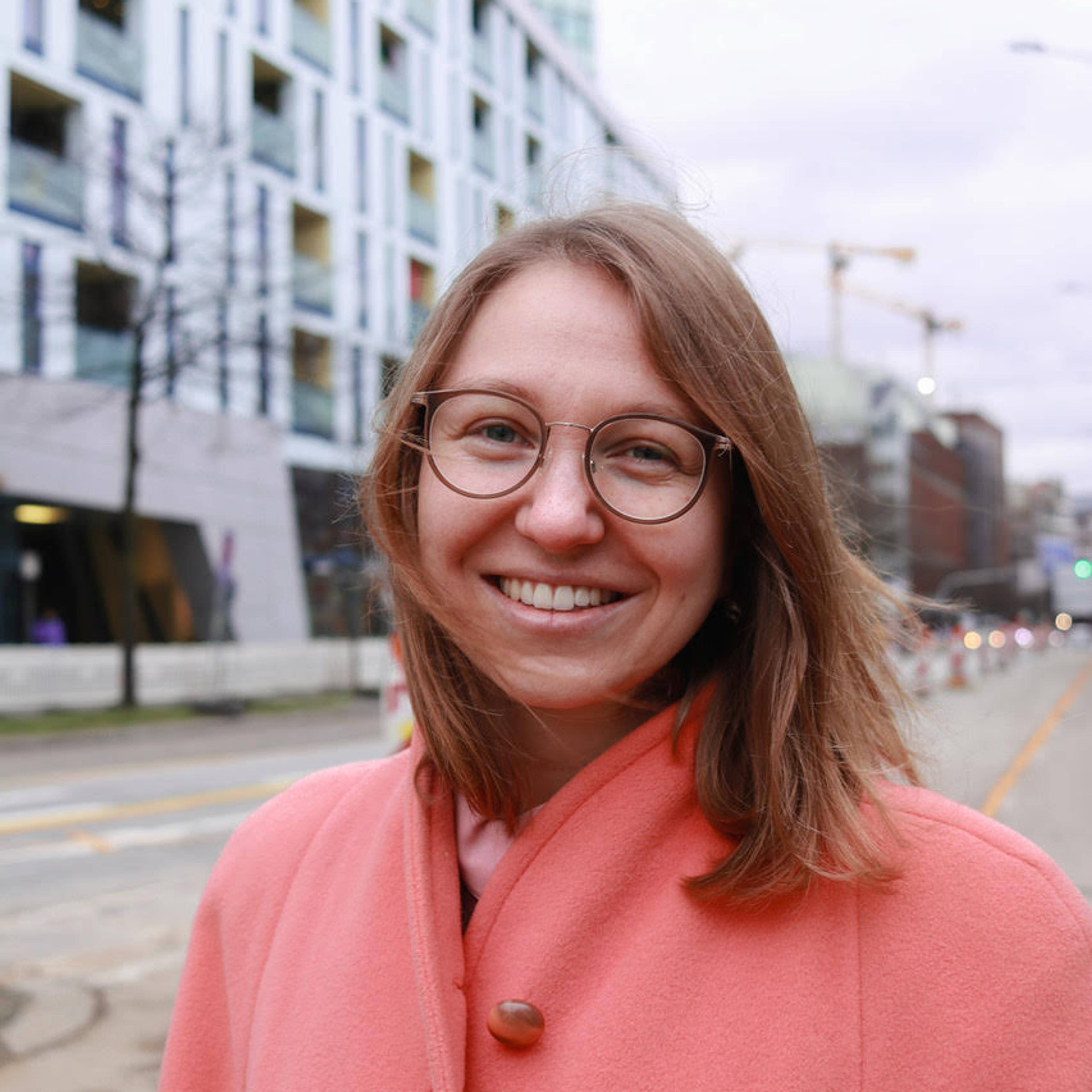 A person wearing glasses and a coral coat smiles at the camera. They stand on a city street with modern buildings and construction cranes in the background. Its an overcast day.