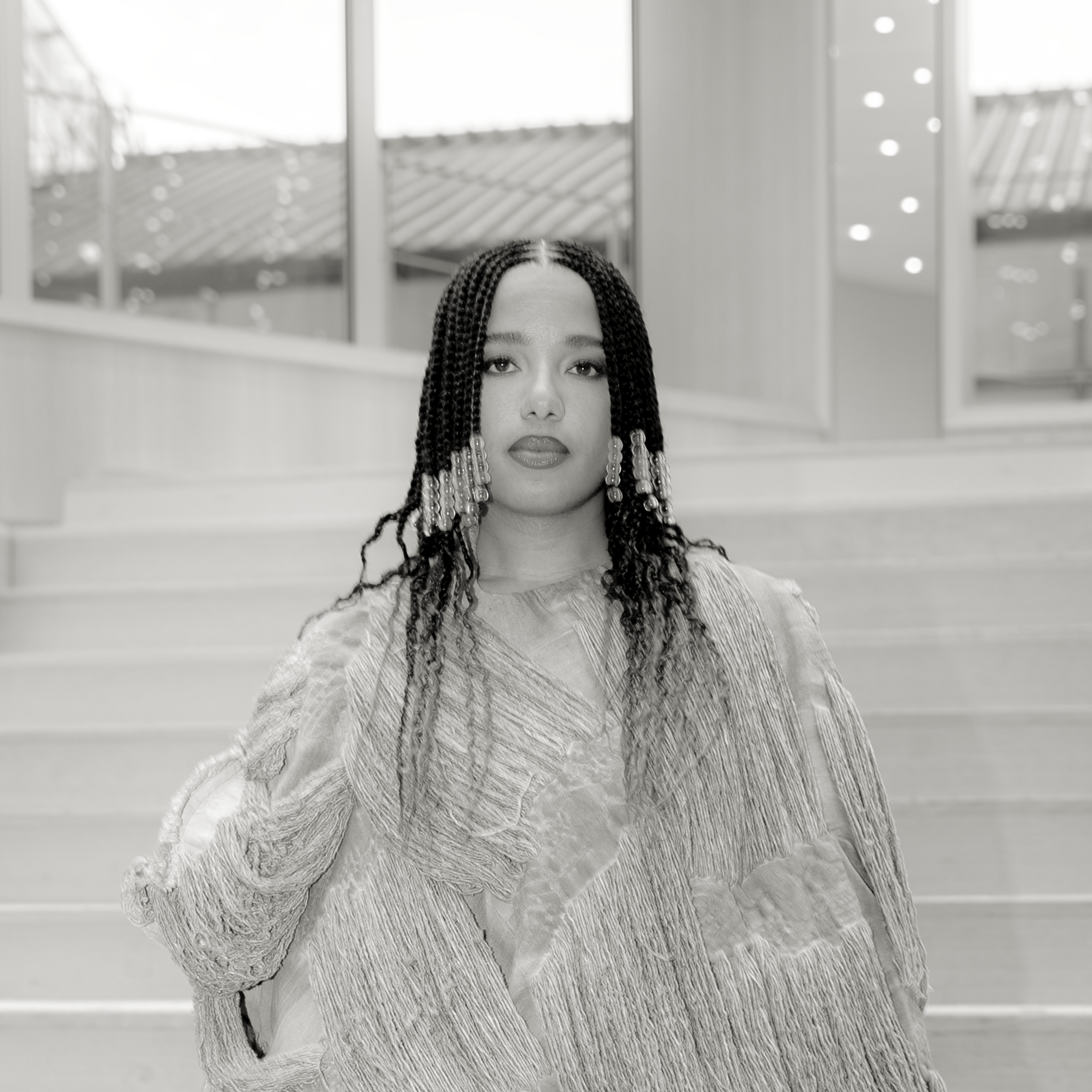 A person with braided hair and decorative beads stands on steps, wearing a textured, flowing outfit. The background features a modern interior with large windows and ceiling lights.