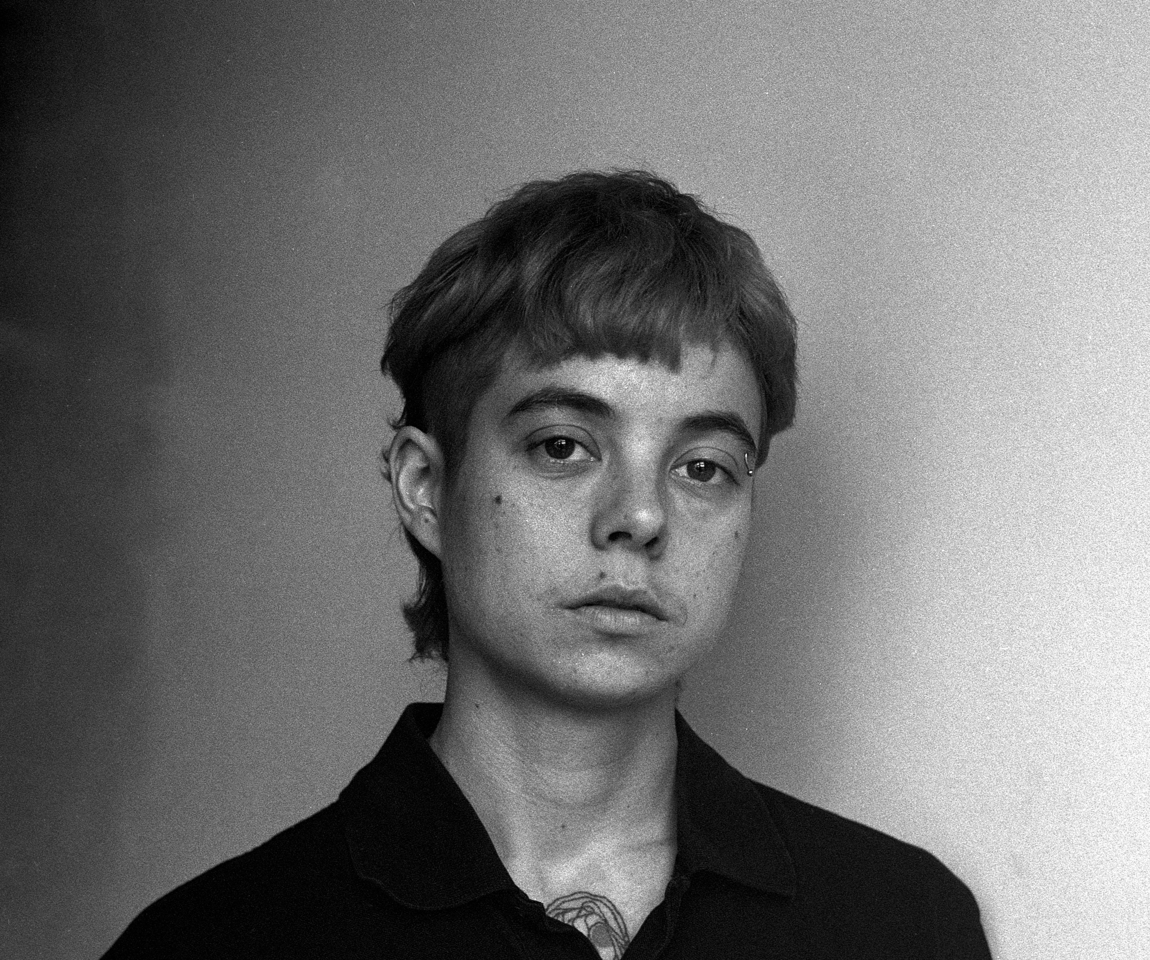 Black and white portrait of a person with short hair, looking at the camera with a neutral expression. They are wearing a dark collared shirt and have a tattoo visible on their neck. The background is plain.