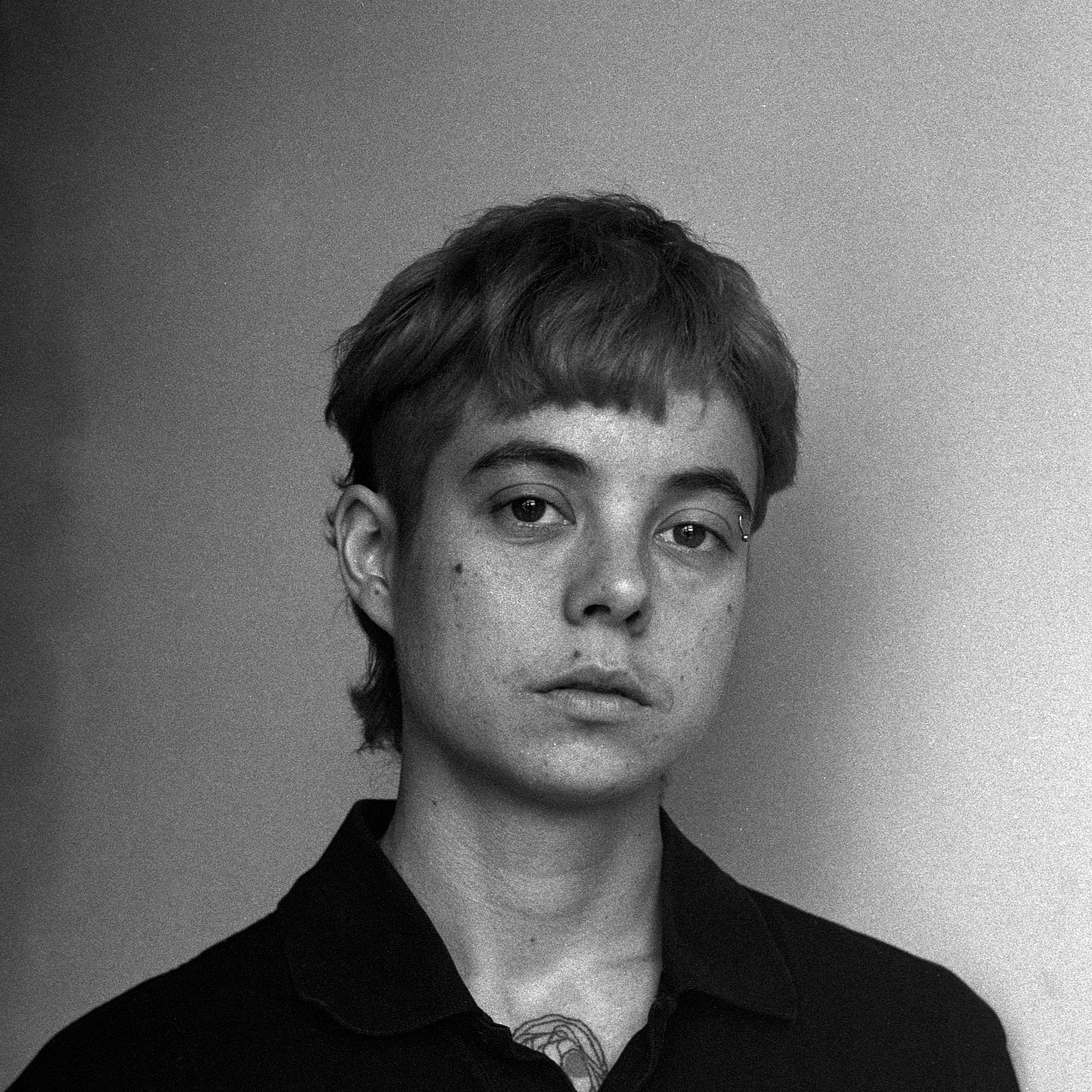 Black and white portrait of a person with short hair, looking at the camera with a neutral expression. They are wearing a dark collared shirt and have a tattoo visible on their neck. The background is plain.