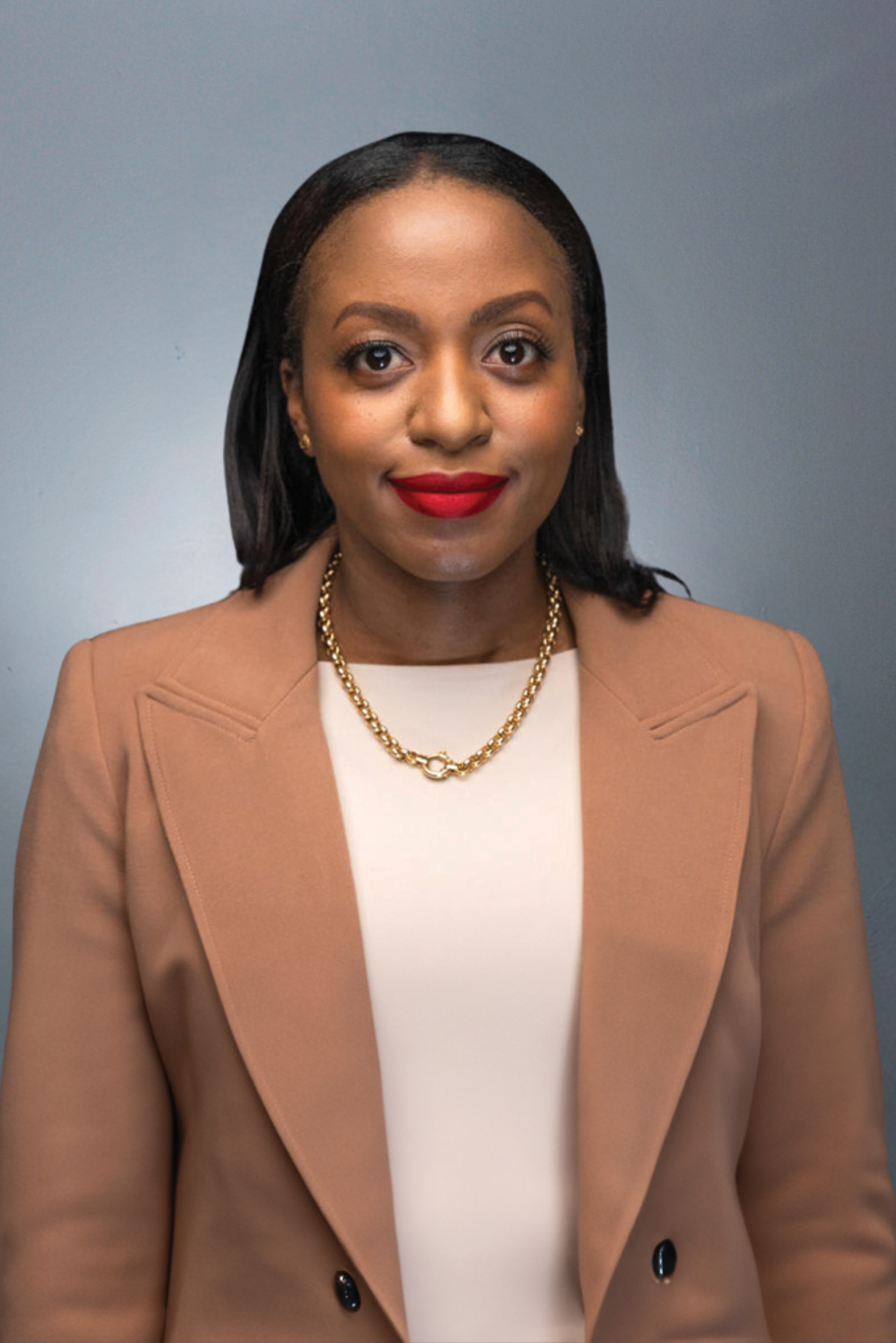 A person with long dark hair wearing a beige blazer, light top, and gold necklace looks at the camera against a neutral grey background.