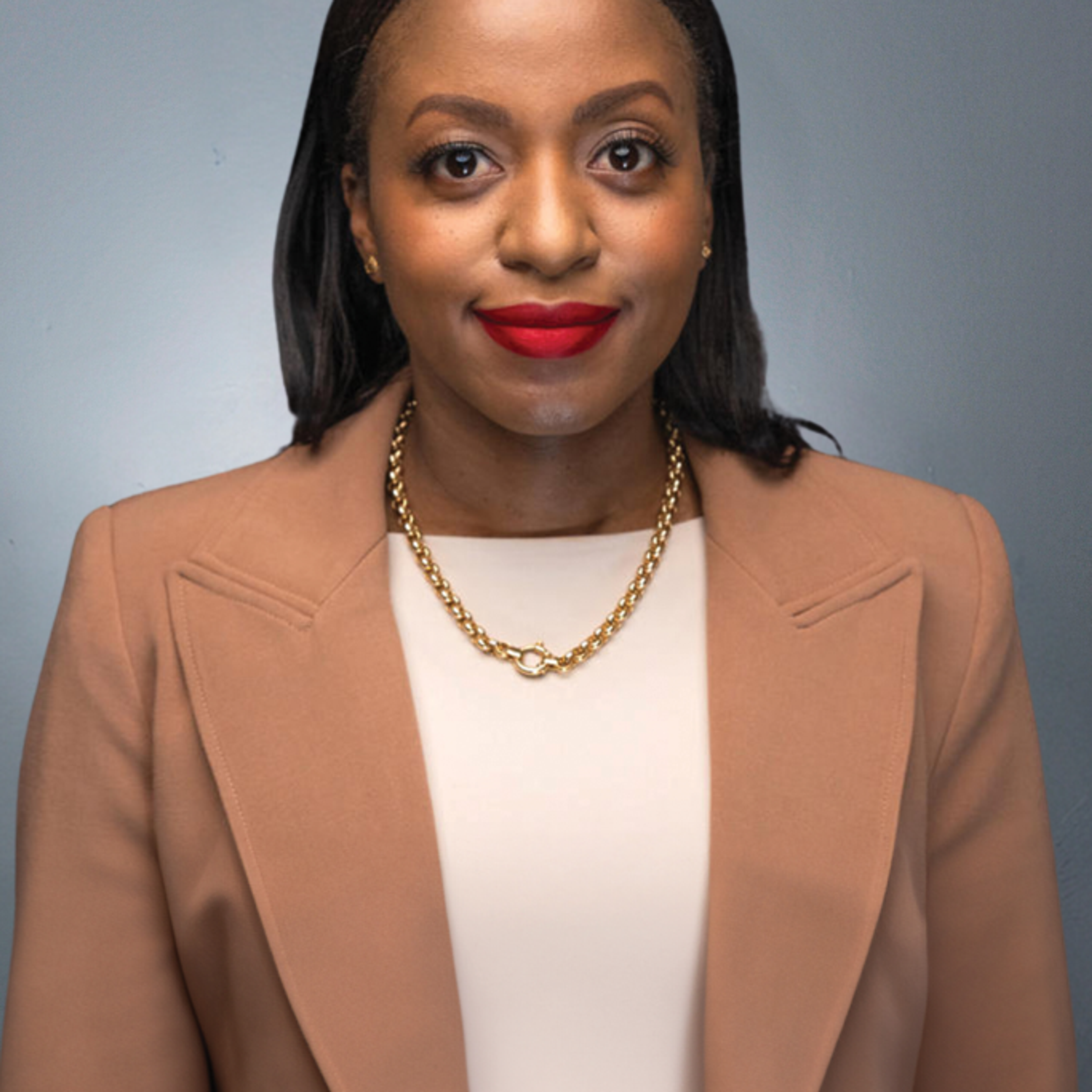 A person with long dark hair wearing a beige blazer, light top, and gold necklace looks at the camera against a neutral grey background.