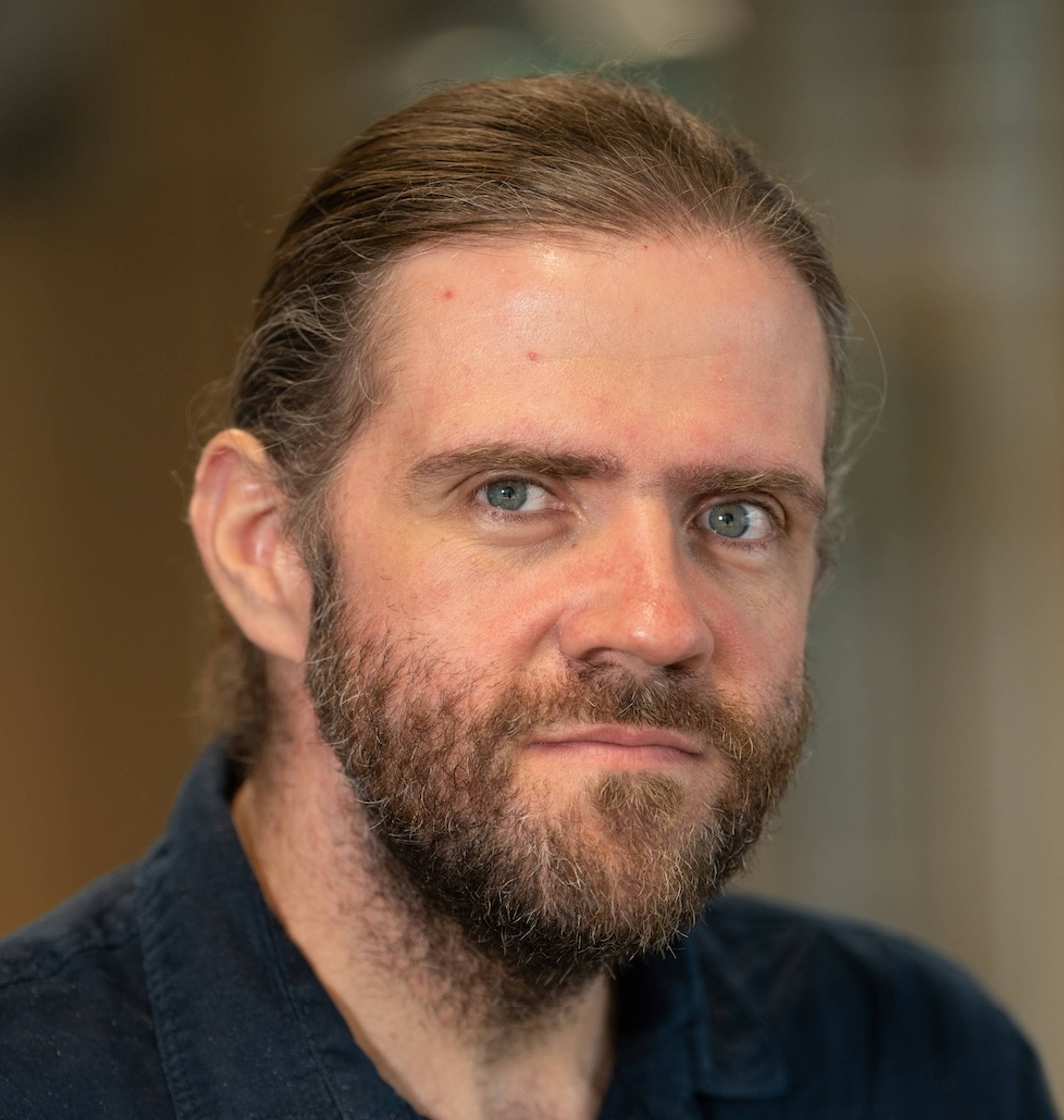 Portrait of a thoughtful-looking man with a beard and long, tied-back hair against a blurred background.
