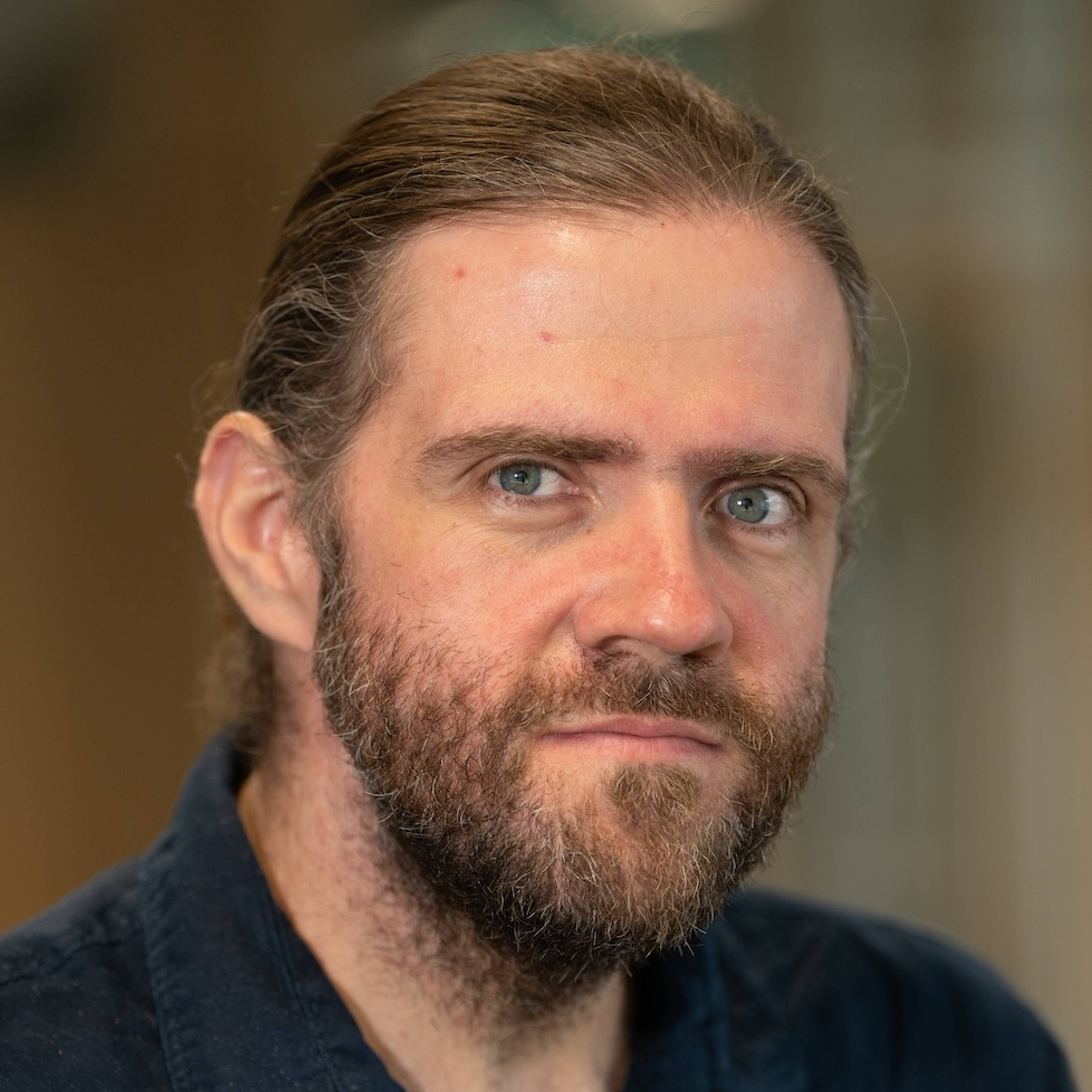 Portrait of a thoughtful-looking man with a beard and long, tied-back hair against a blurred background.