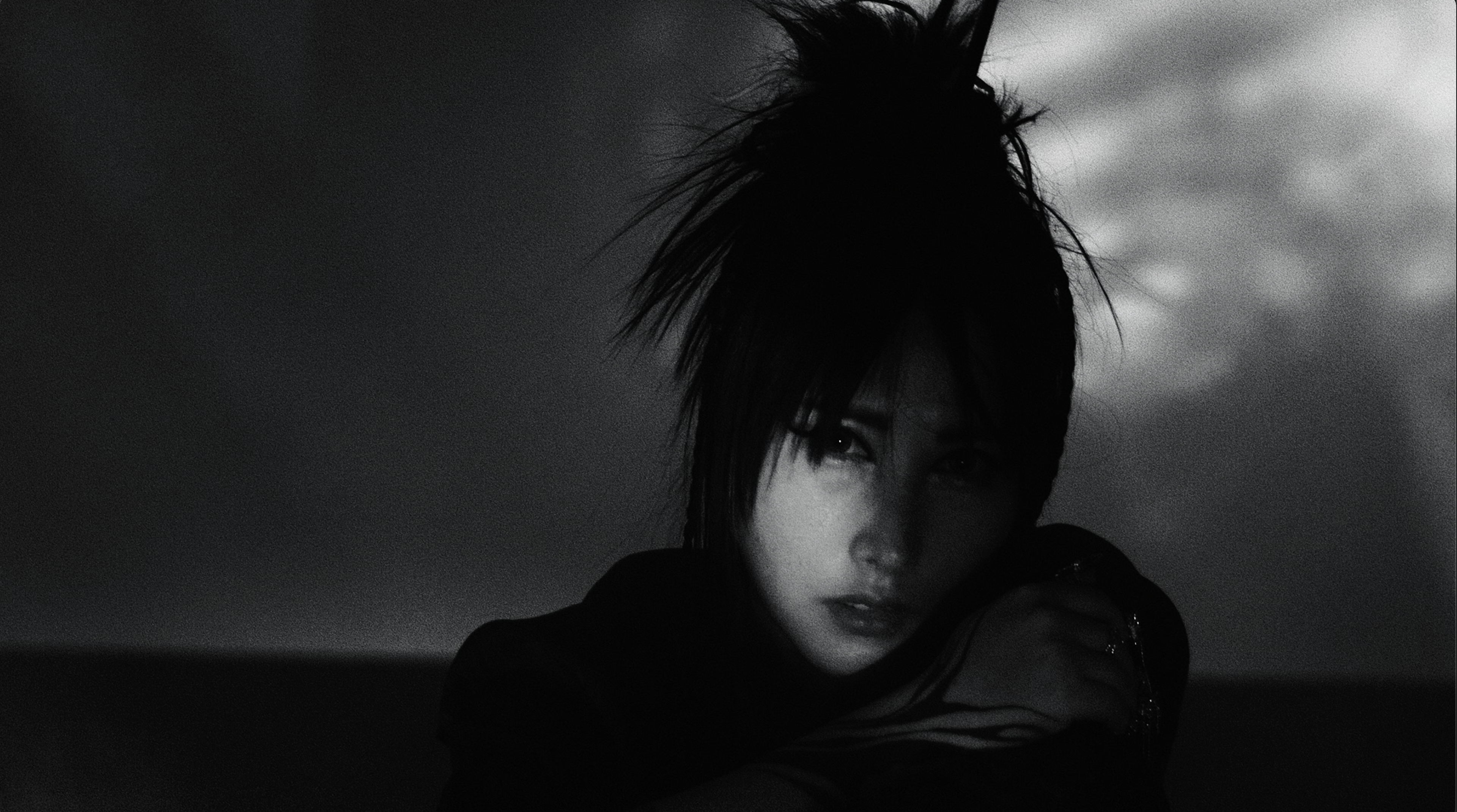 A black and white portrait of a person with dark, tousled hair, looking directly at the camera with a neutral expression. They are partially shadowed, creating a dramatic and moody atmosphere.