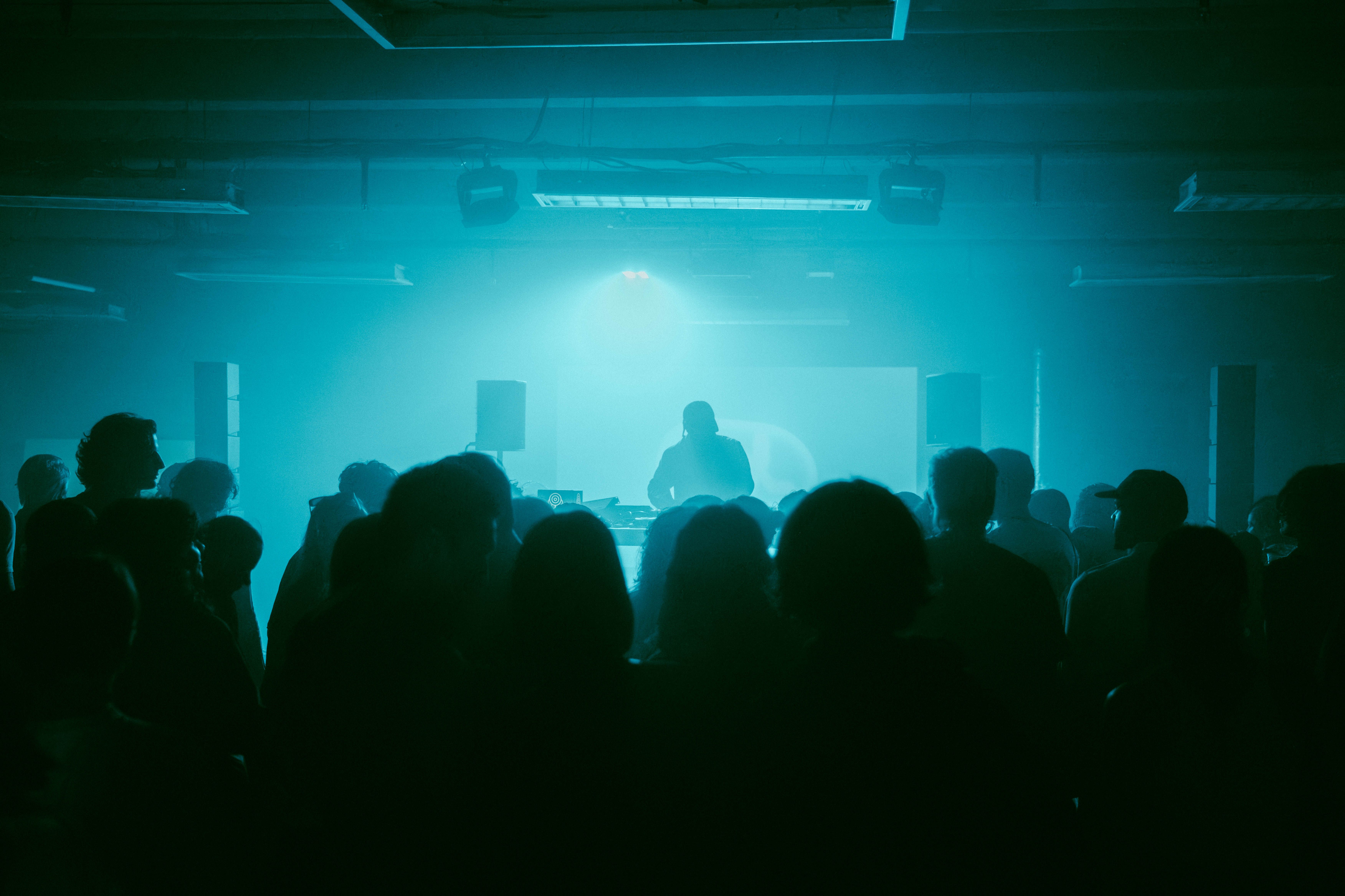 Publikum in einem dunklen Raum während der CEL Launch Night im CANK Berlin, Neukölln. Im Vordergrund die Silhouetten der Besucher:innen, auf der Bühne DJ Bobby Beethoven im blau-grünen Licht.