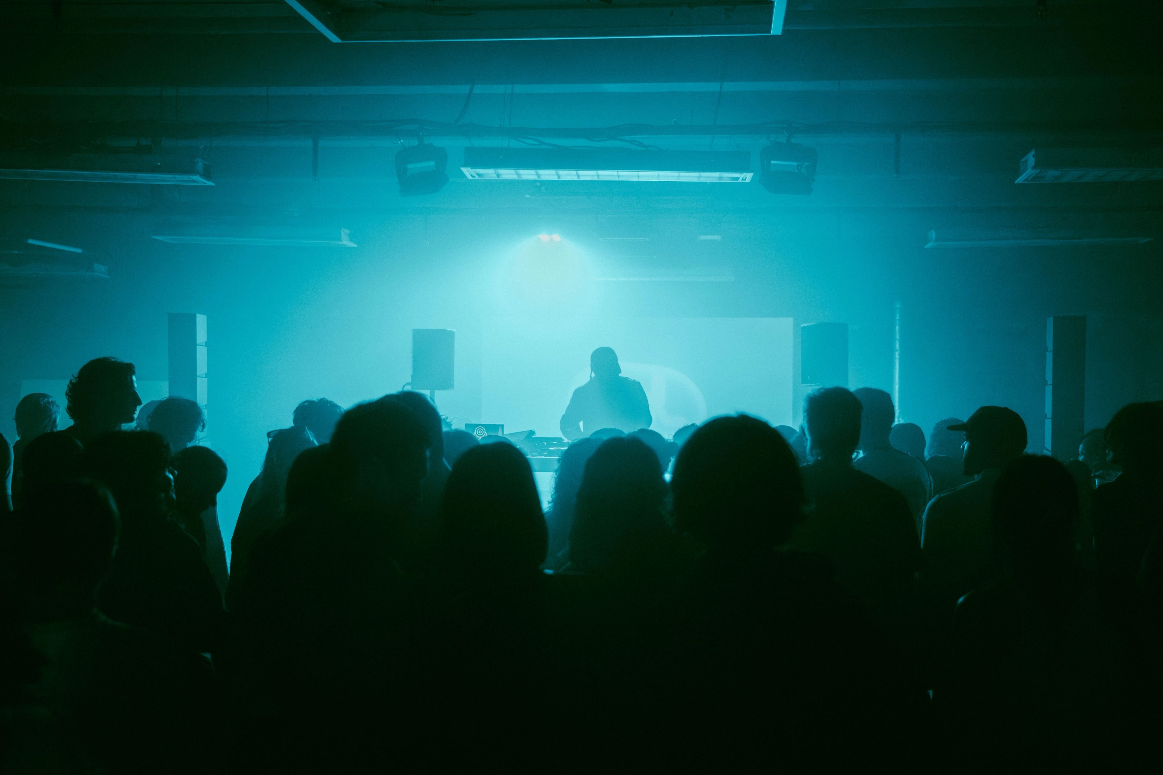 A DJ performs in front of a crowd in a room dimly lit with blue light.
