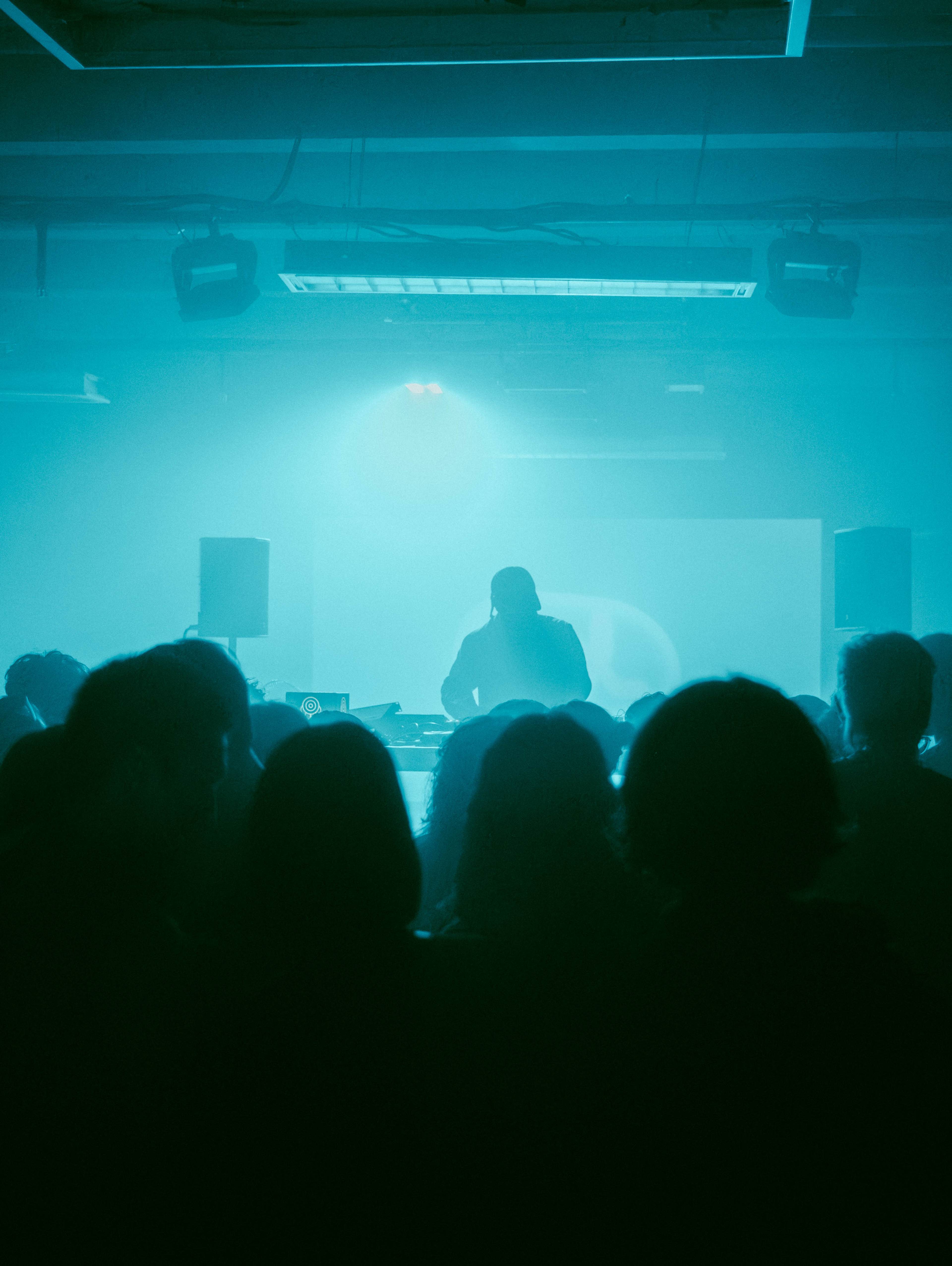 Publikum in einem dunklen Raum während der CEL Launch Night im CANK Berlin, Neukölln. Im Vordergrund die Silhouetten der Besucher:innen, auf der Bühne DJ Bobby Beethoven im blau-grünen Licht.