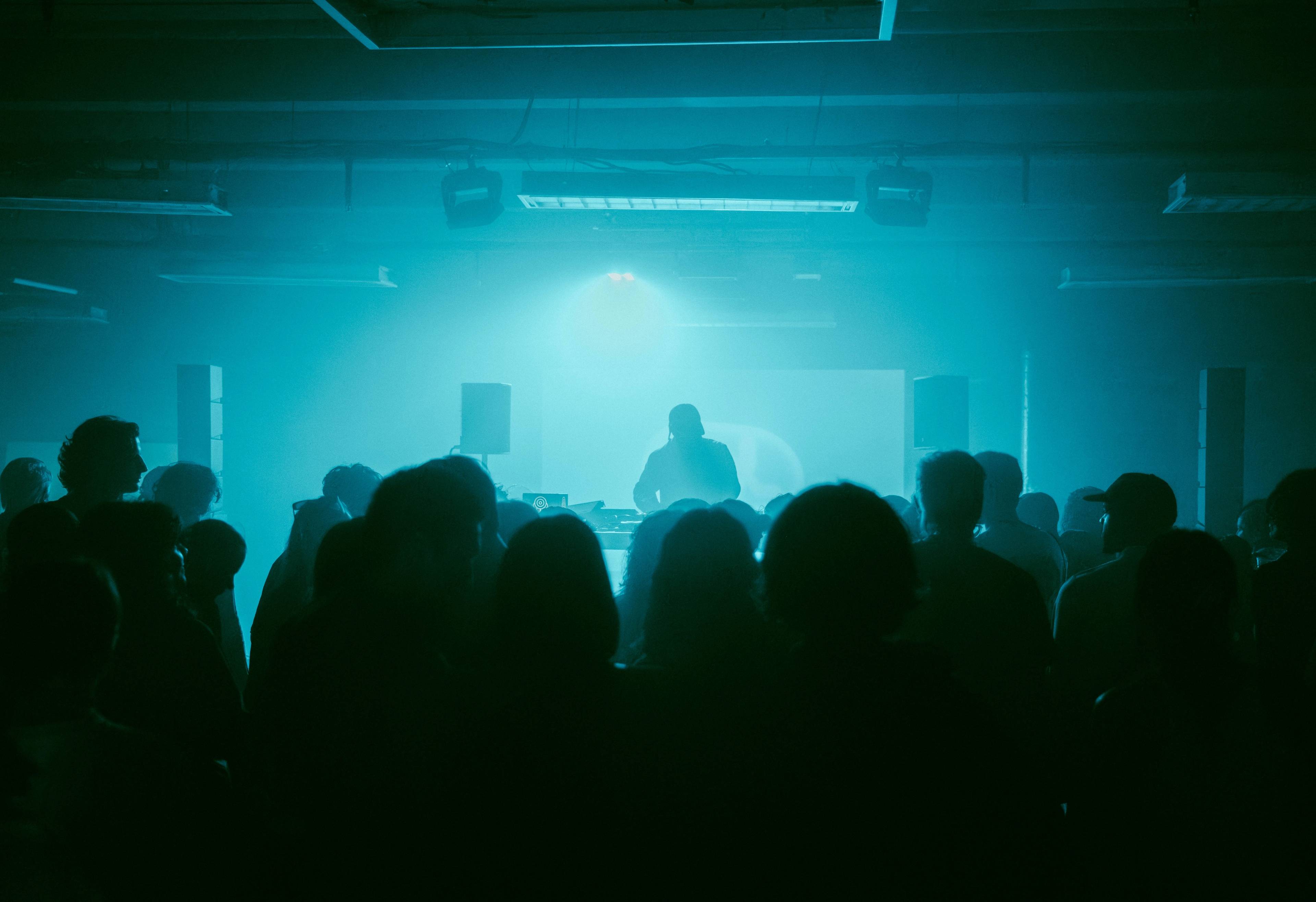 Publikum in einem dunklen Raum während der CEL Launch Night im CANK Berlin, Neukölln. Im Vordergrund die Silhouetten der Besucher:innen, auf der Bühne DJ Bobby Beethoven im blau-grünen Licht.
