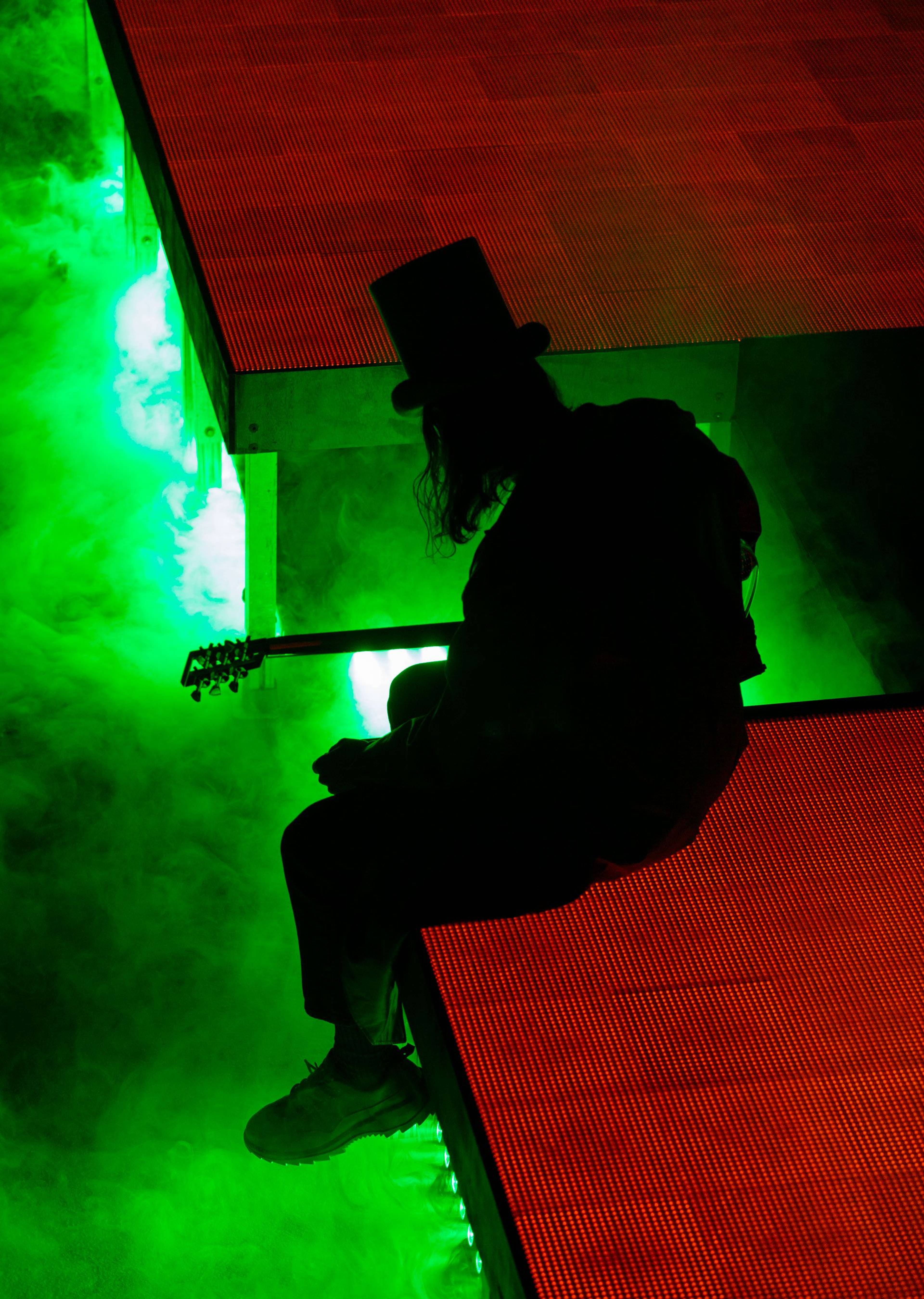 Member of music duo Amnesia Scanner sitting on a stage ledge and playing guitar above a field of green fog.