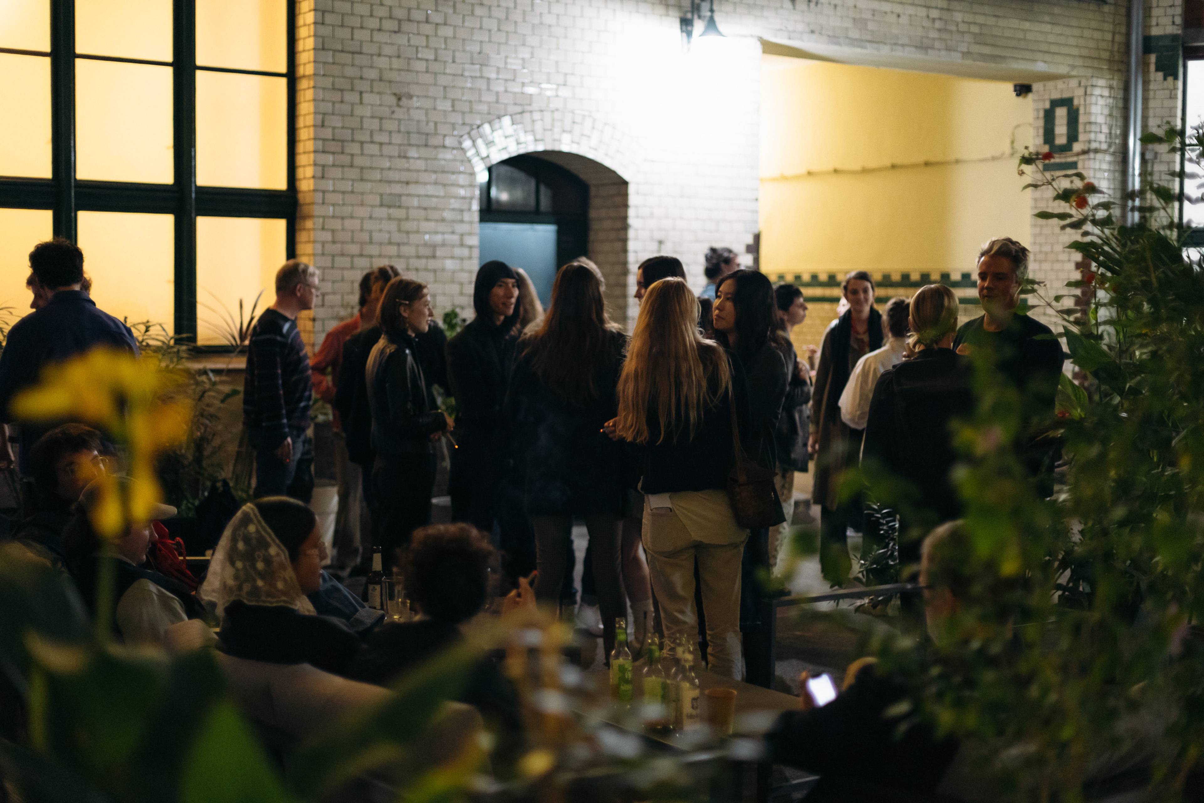 Menschen stehen nach einem Gespräch zusammen und unterhalten sich in einem hellen Innenhof mit weißen Fliesenwänden, großen Fenstern und vielen Pflanzen. Im Vordergrund sitzen weitere Besucher:innen mit Getränken an Tischen und setzen die Gespräche nach dem Künstler:innengespräch fort.