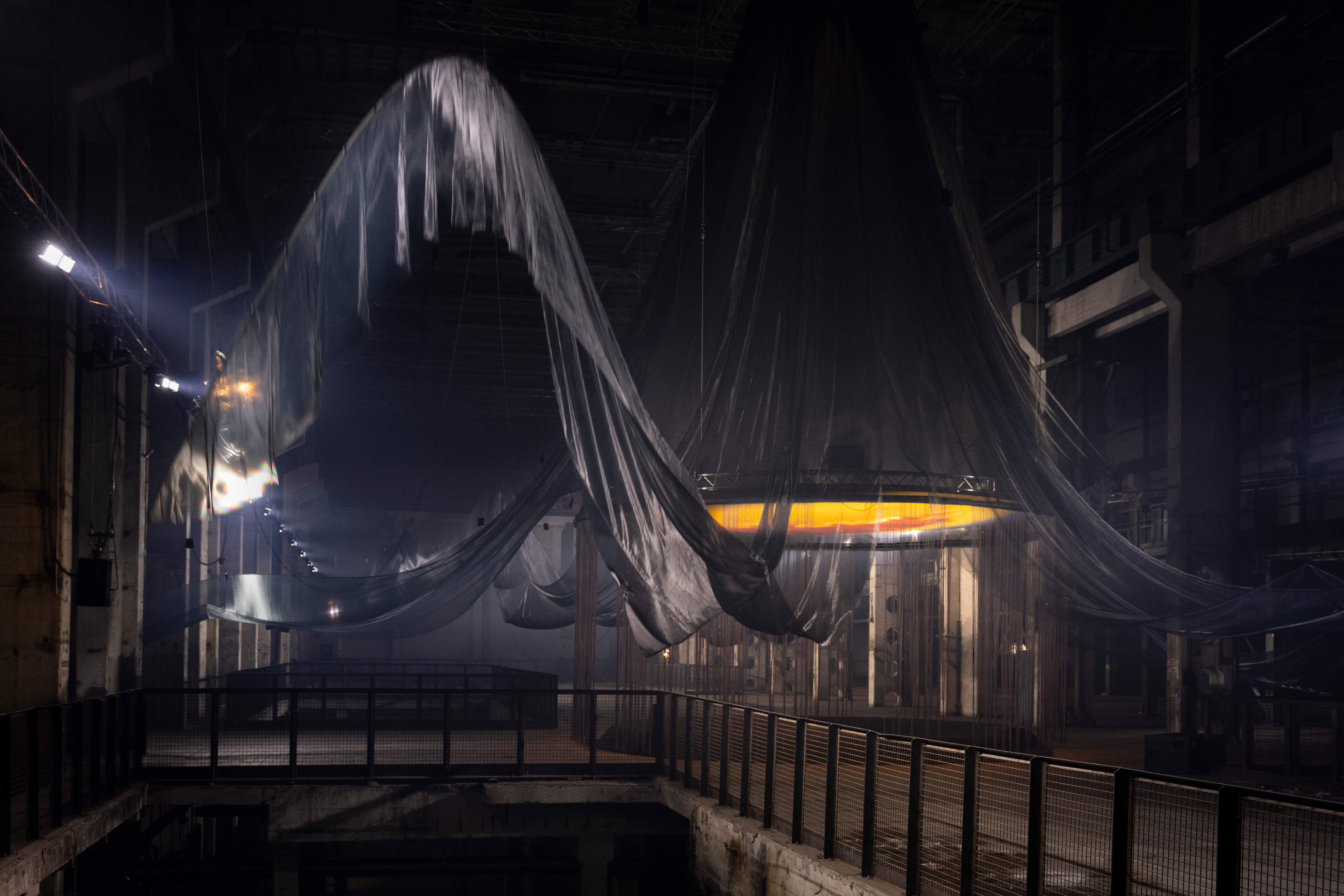 Eine großflächige, immersive Kunstinstallation in einer industriellen Umgebung, mit fließendem, metallisch glänzendem Stoff, der sich durch den Raum zieht. Die gedämpfte Beleuchtung wird von kühlem blauen Licht durchbrochen, das die Textur des Stoffes hervorhebt, während er von der Decke herabfällt. Die Szene wirkt ätherisch und fast surreal, wobei der Stoff scheinbar schwerelos schwebt. Der Raum ist geprägt von Betonwänden, Metallträgern und einer hohen, weitläufigen Decke, was das Gefühl von Weite und Tiefe verstärkt.