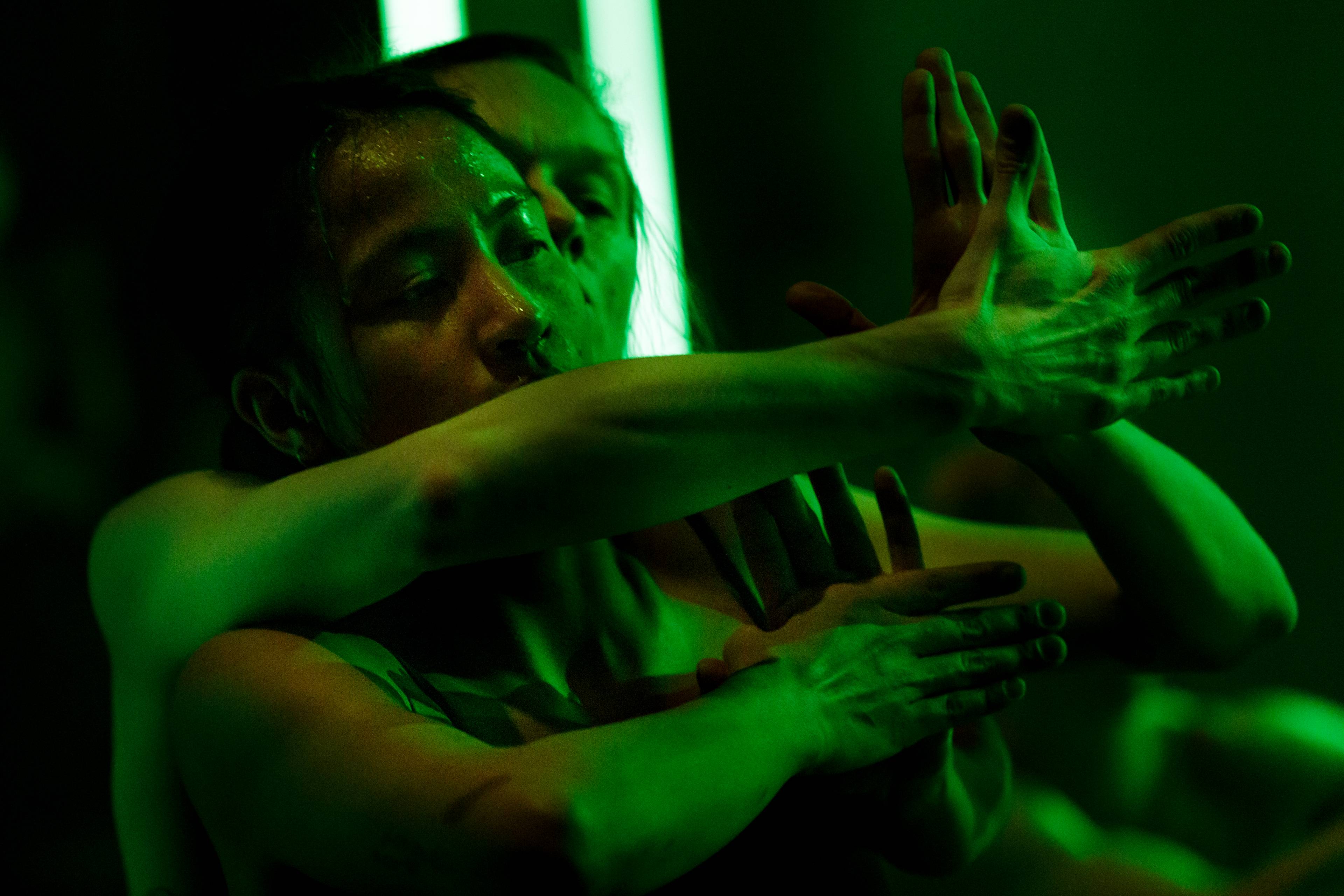 Dancer bathed in green light embracing a fellow performer from behind and performing a coordinated hand gesture.