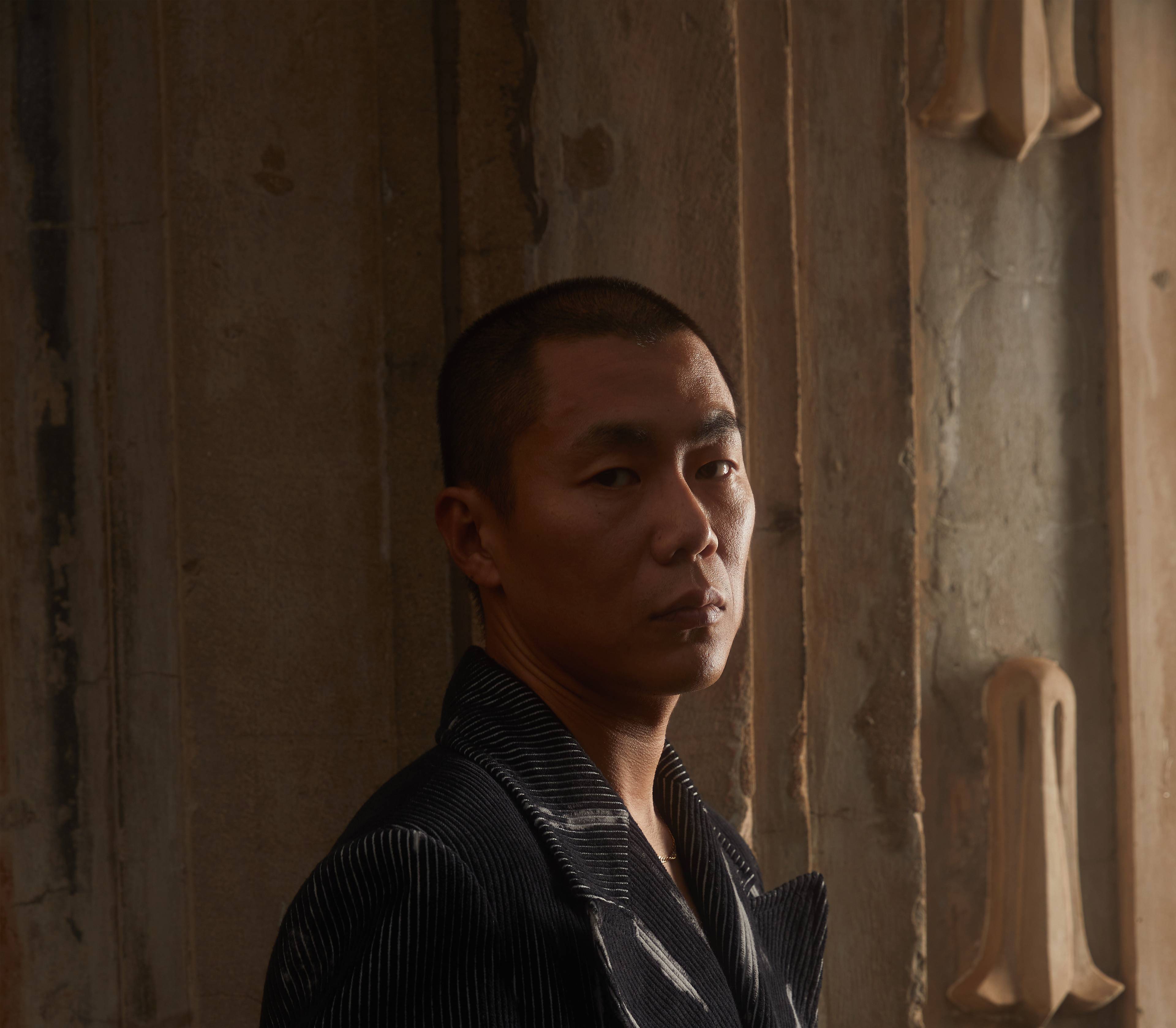 A person with short hair and a striped jacket looks seriously into the camera, standing in front of a textured wall bathed in warm, side lighting.