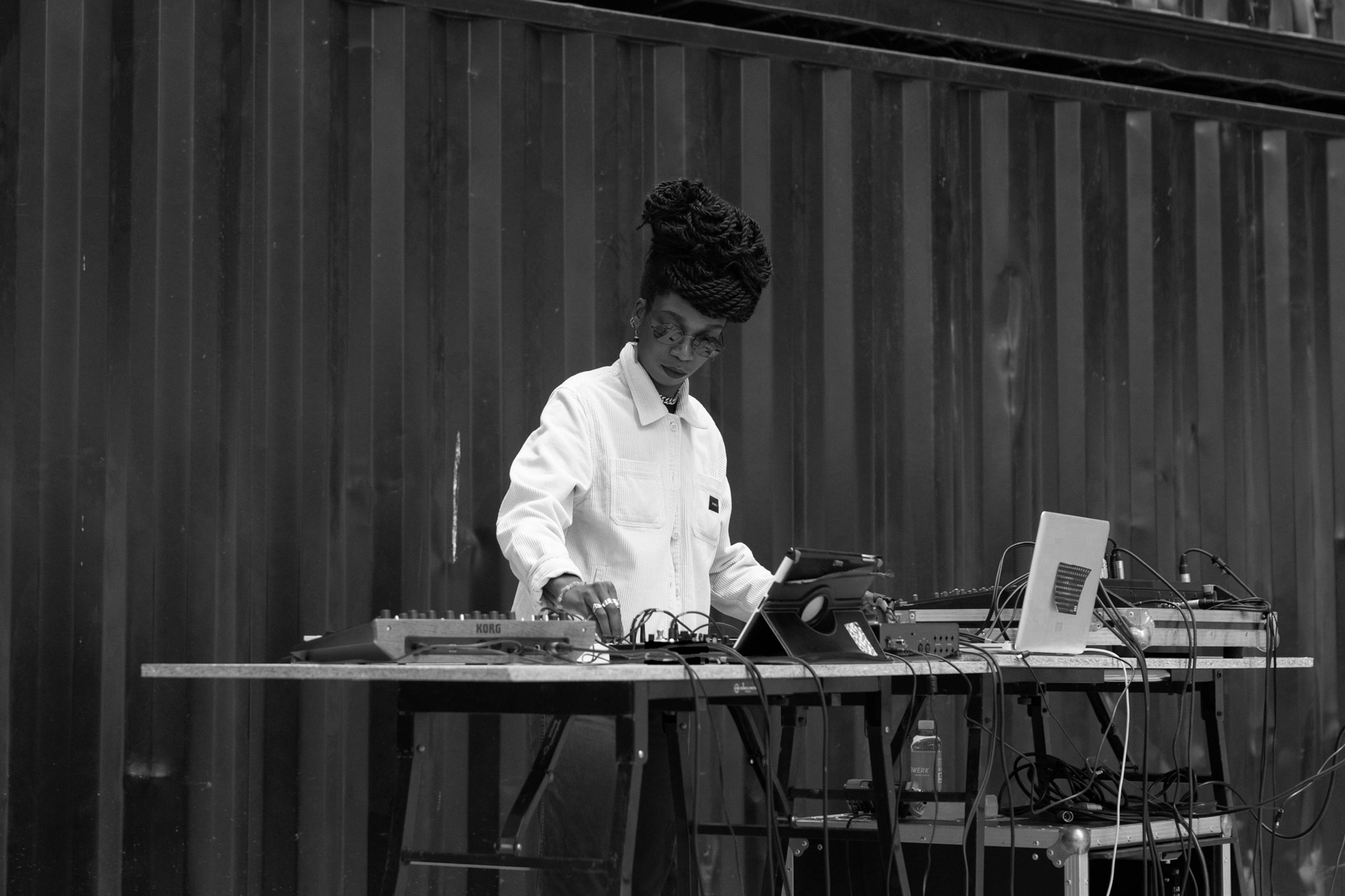 A person with braided hair is standing at a table with electronic equipment, wearing a white jacket and adjusting the controls. The setup includes a laptop and various audio devices against a dark, vertical panel backdrop.