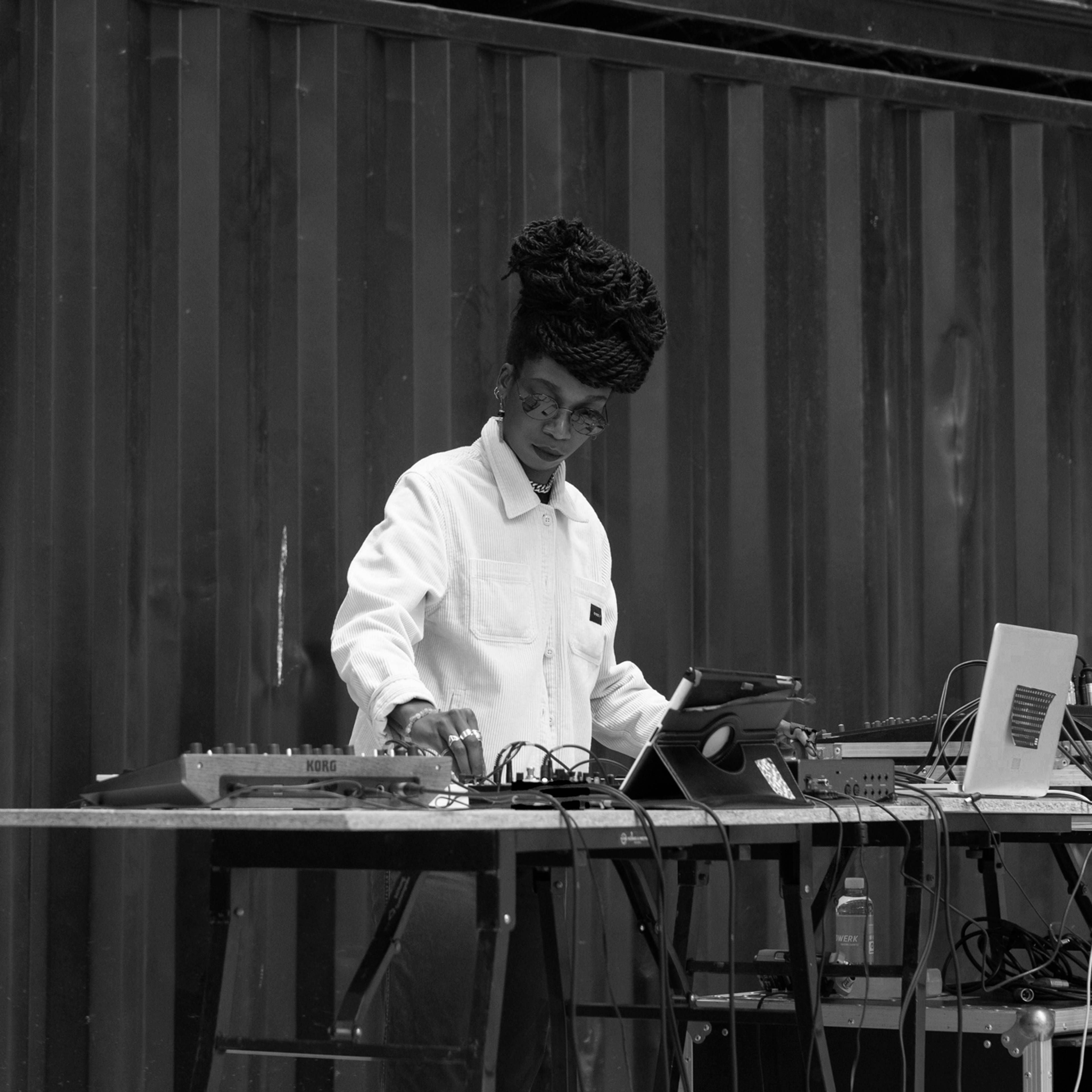 A person with braided hair is standing at a table with electronic equipment, wearing a white jacket and adjusting the controls. The setup includes a laptop and various audio devices against a dark, vertical panel backdrop.
