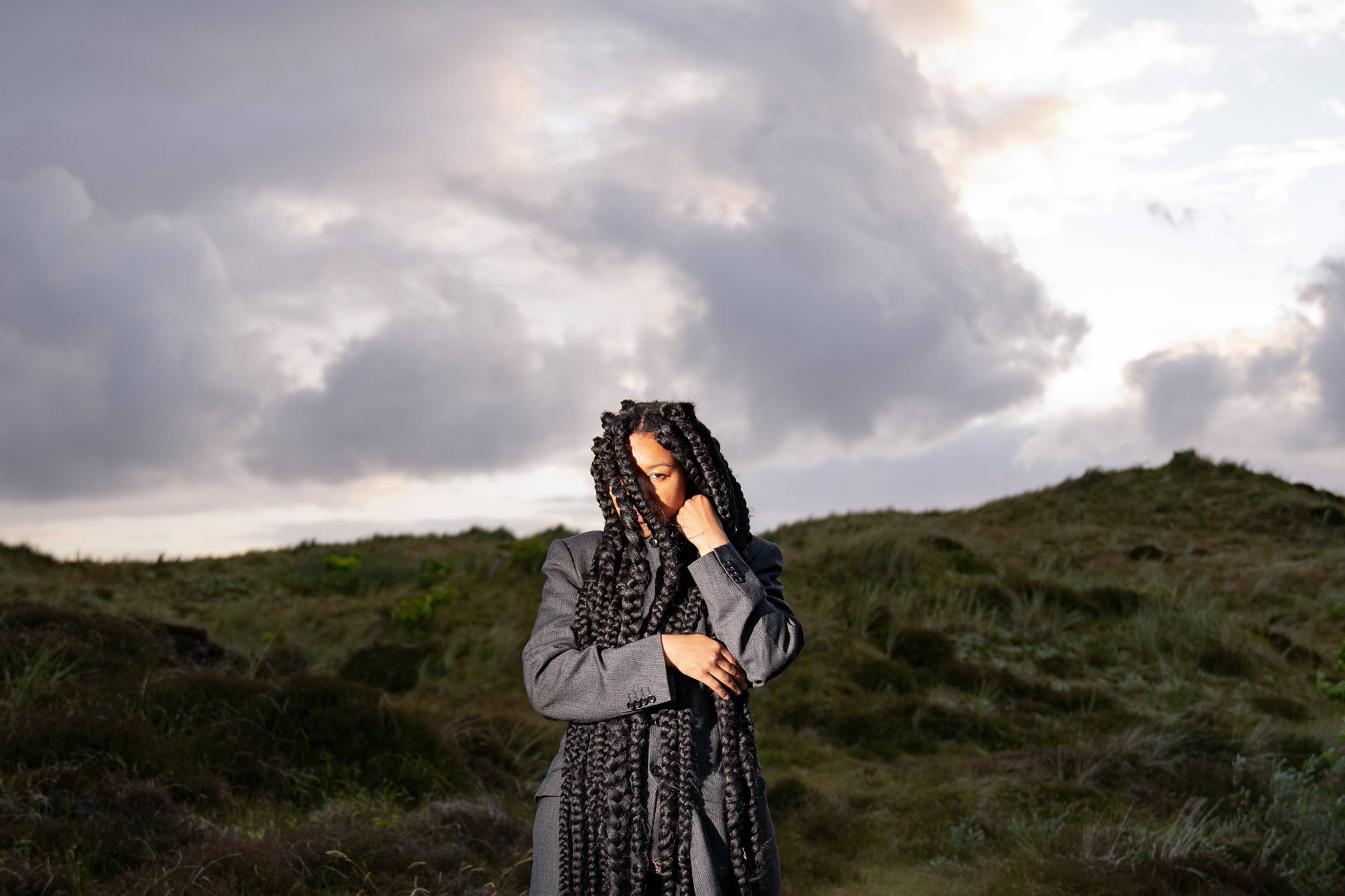 A person with long braided hair and a gray coat stands outdoors, looking contemplative. The background features a hilly landscape and a dramatic, cloudy sky.