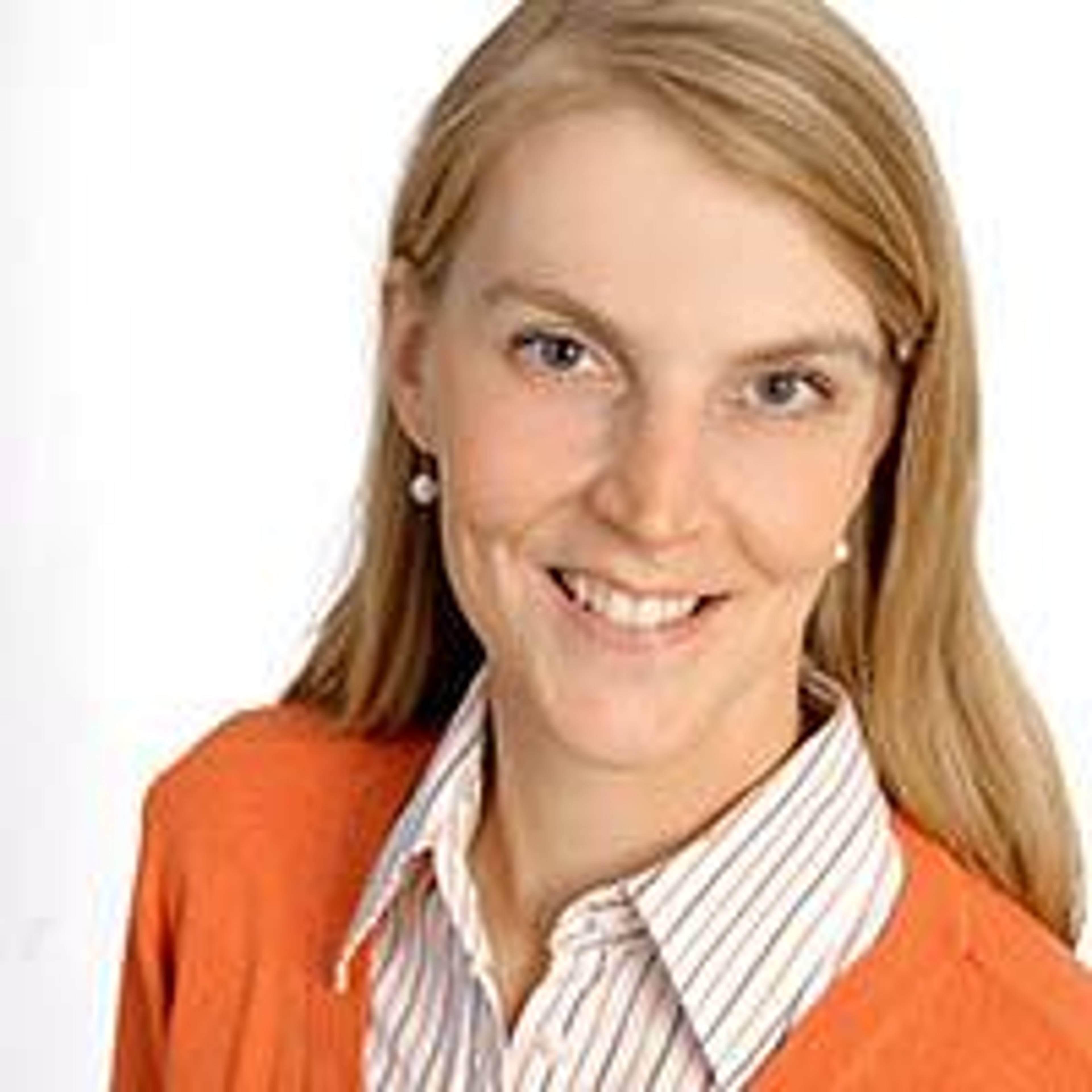 Smiling person with long blonde hair, wearing an orange sweater over a striped shirt, against a plain white background.