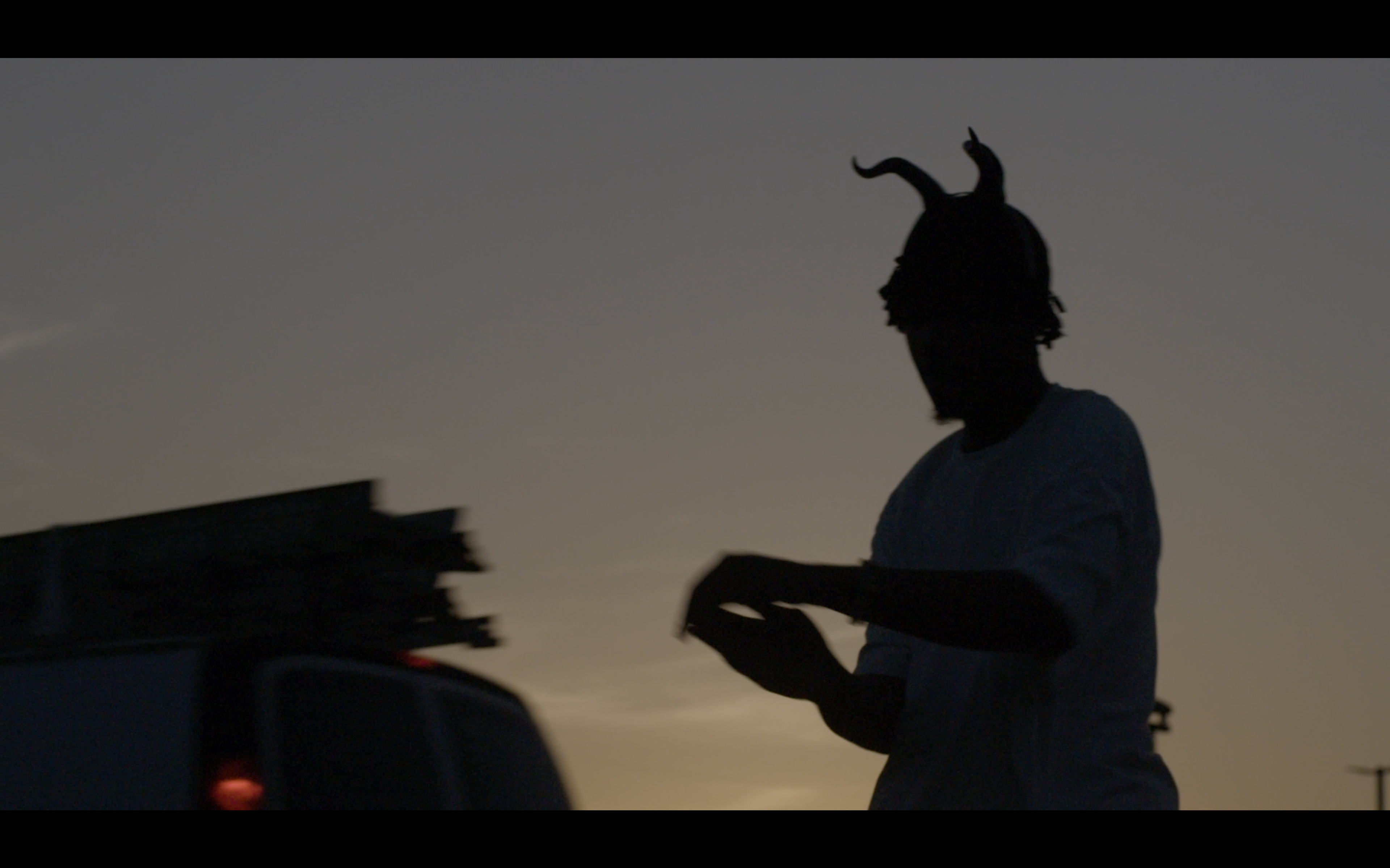 Silhouette einer Person mit einem Kopfschmuck aus gebogenen Hörnern, die bei Sonnenuntergang neben einem Fahrzeug steht.
