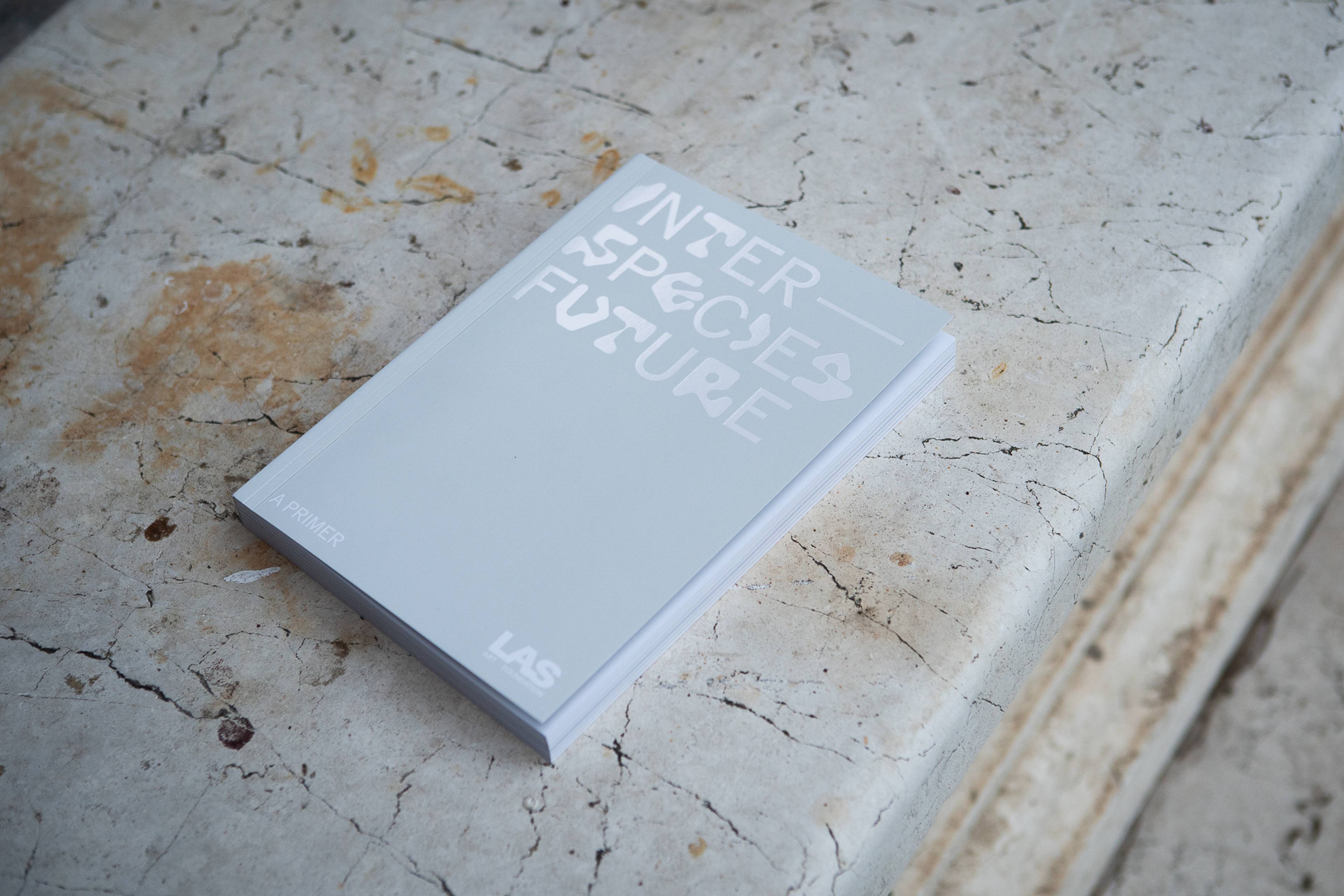 Ein Buch mit dem Titel Interspecies Future: A Primer mit einem pastellfarbenen, reflektierenden Einband und stilisiertem Text. Der Hintergrund besteht aus einem Farbverlauf in Grautönen, der ein minimalistisches und modernes Erscheinungsbild vermittelt.
