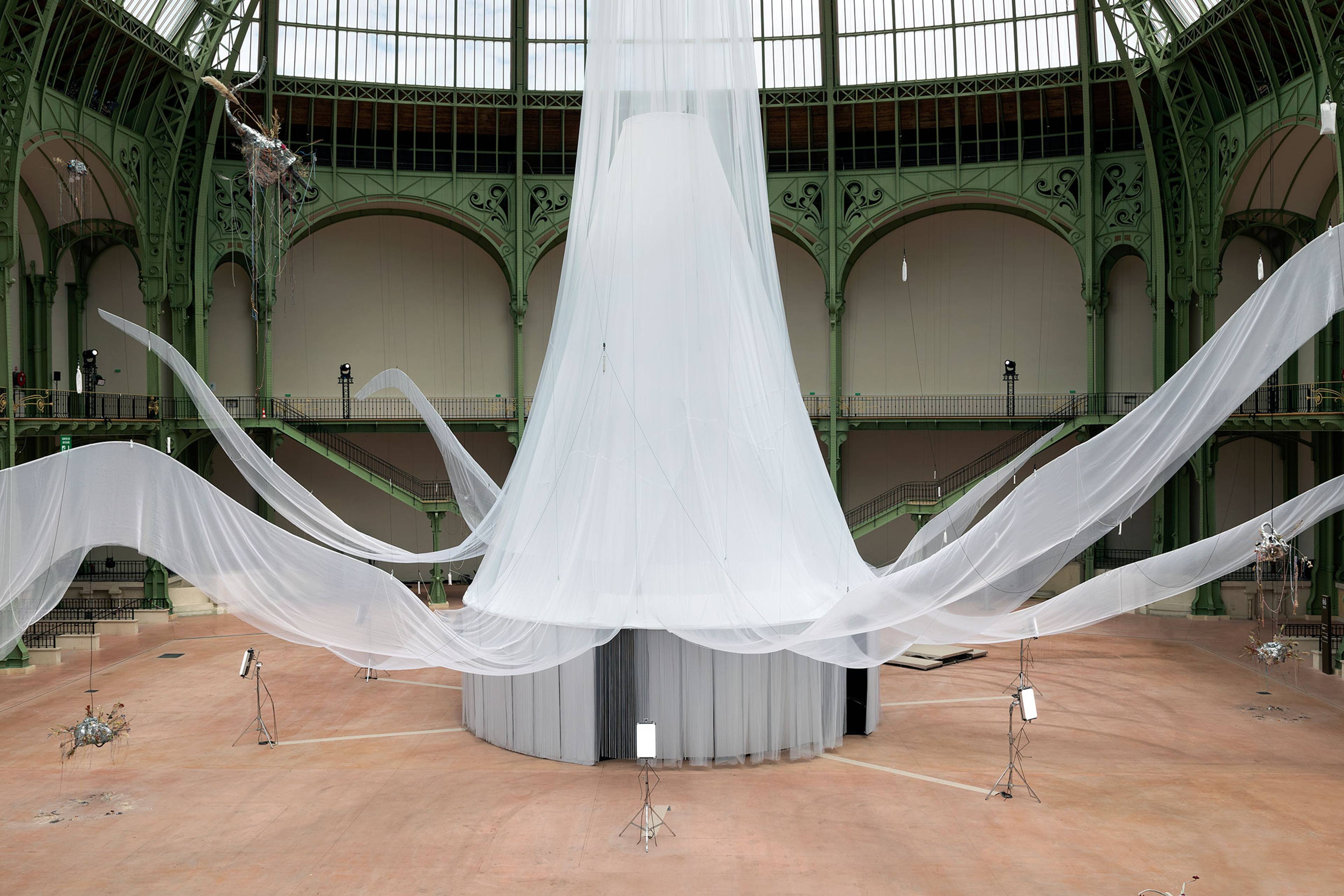 Wide view of Laure Prouvost’s exhibition We Felt A Star Dying at the Grand Palais in Paris, featuring a monumental cylindrical structure draped in translucent white fabric that extends across the historic glass-roofed hall, creating an immersive, dreamlike environment.