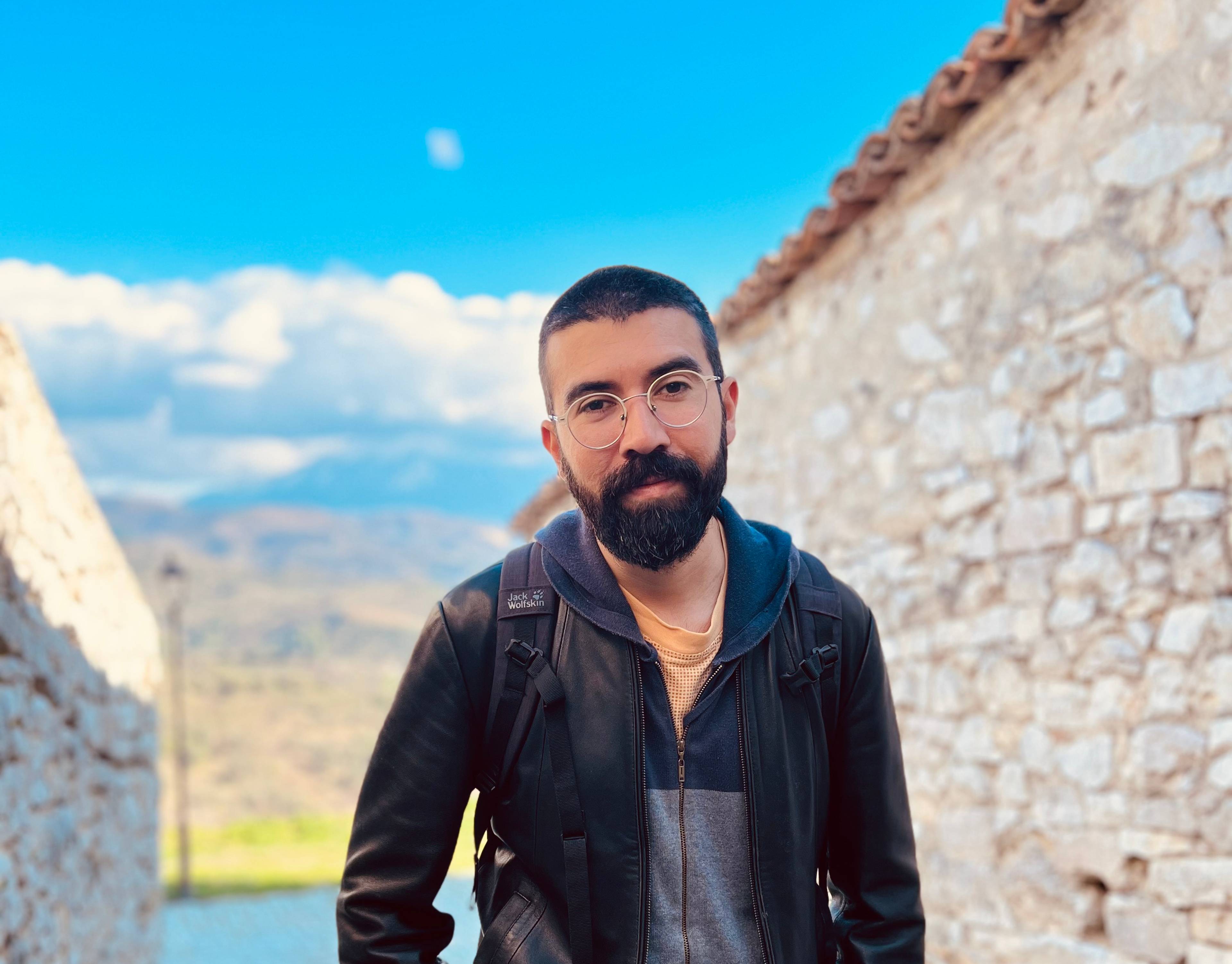 A man with a beard and glasses stands on a cobblestone path between stone walls. He is wearing a leather jacket and a backpack. The sky is clear with some clouds over distant hills.