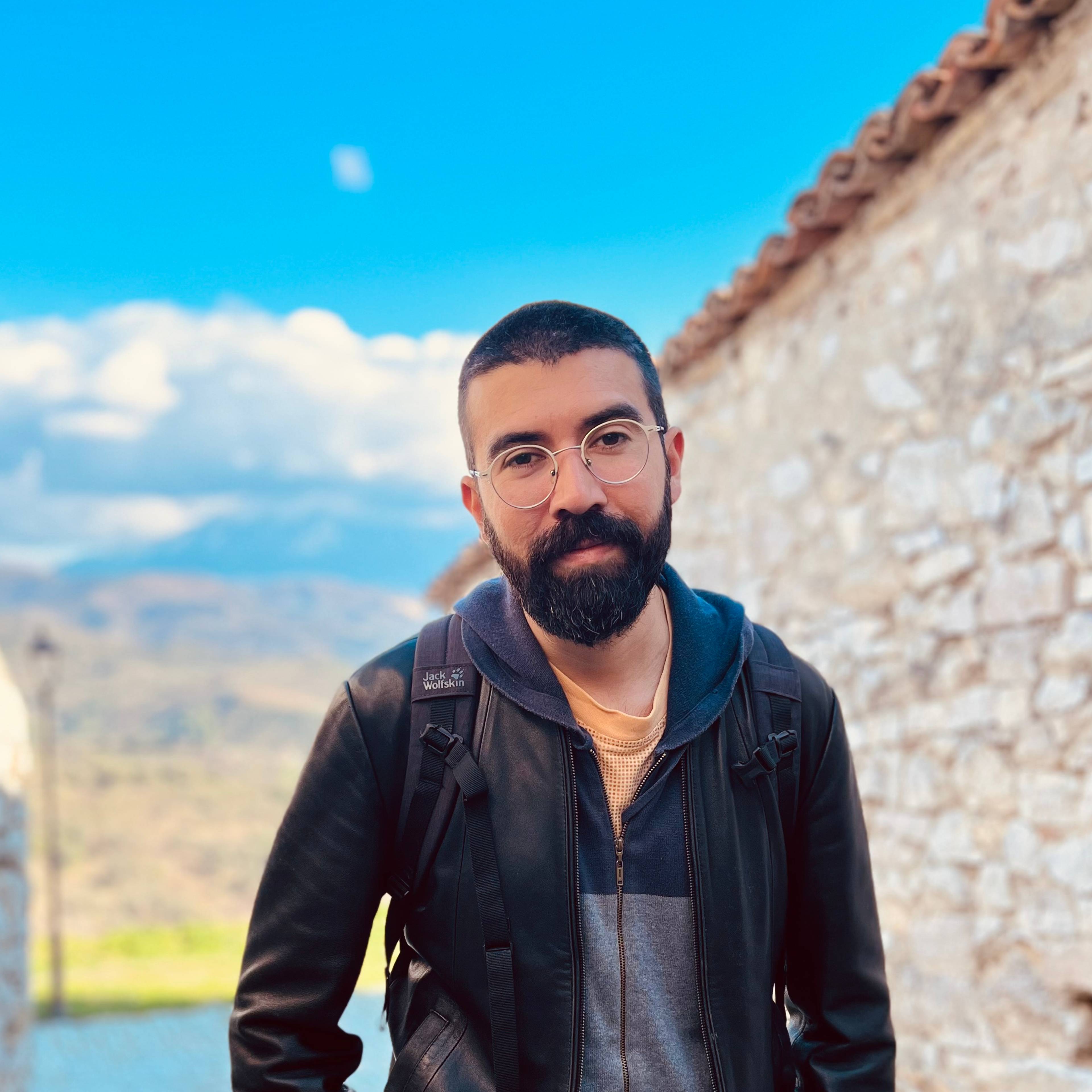 A man with a beard and glasses stands on a cobblestone path between stone walls. He is wearing a leather jacket and a backpack. The sky is clear with some clouds over distant hills.