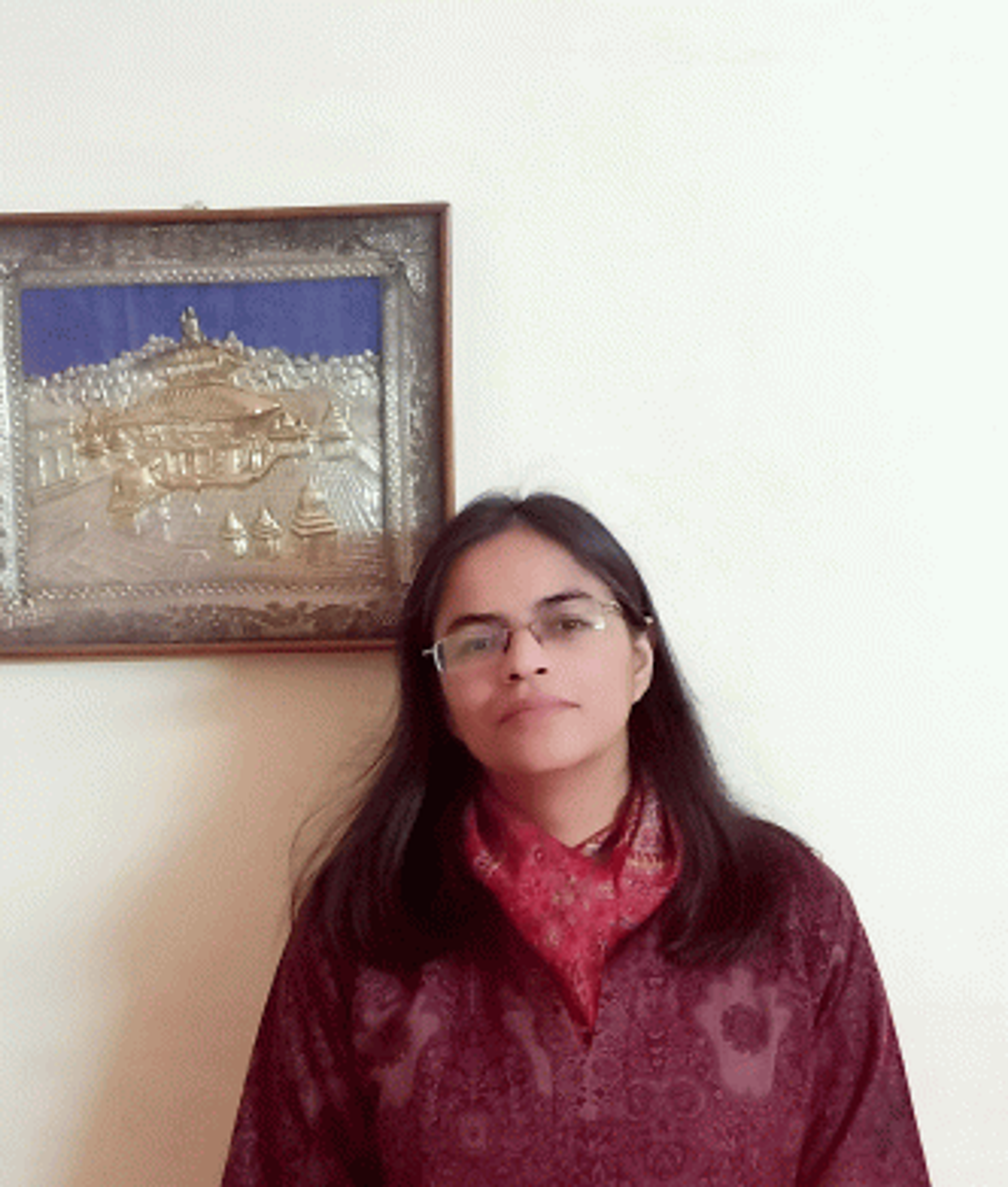 A person with long dark hair and glasses stands indoors, wearing a maroon outfit and a red scarf. A framed artwork is on the wall behind them.