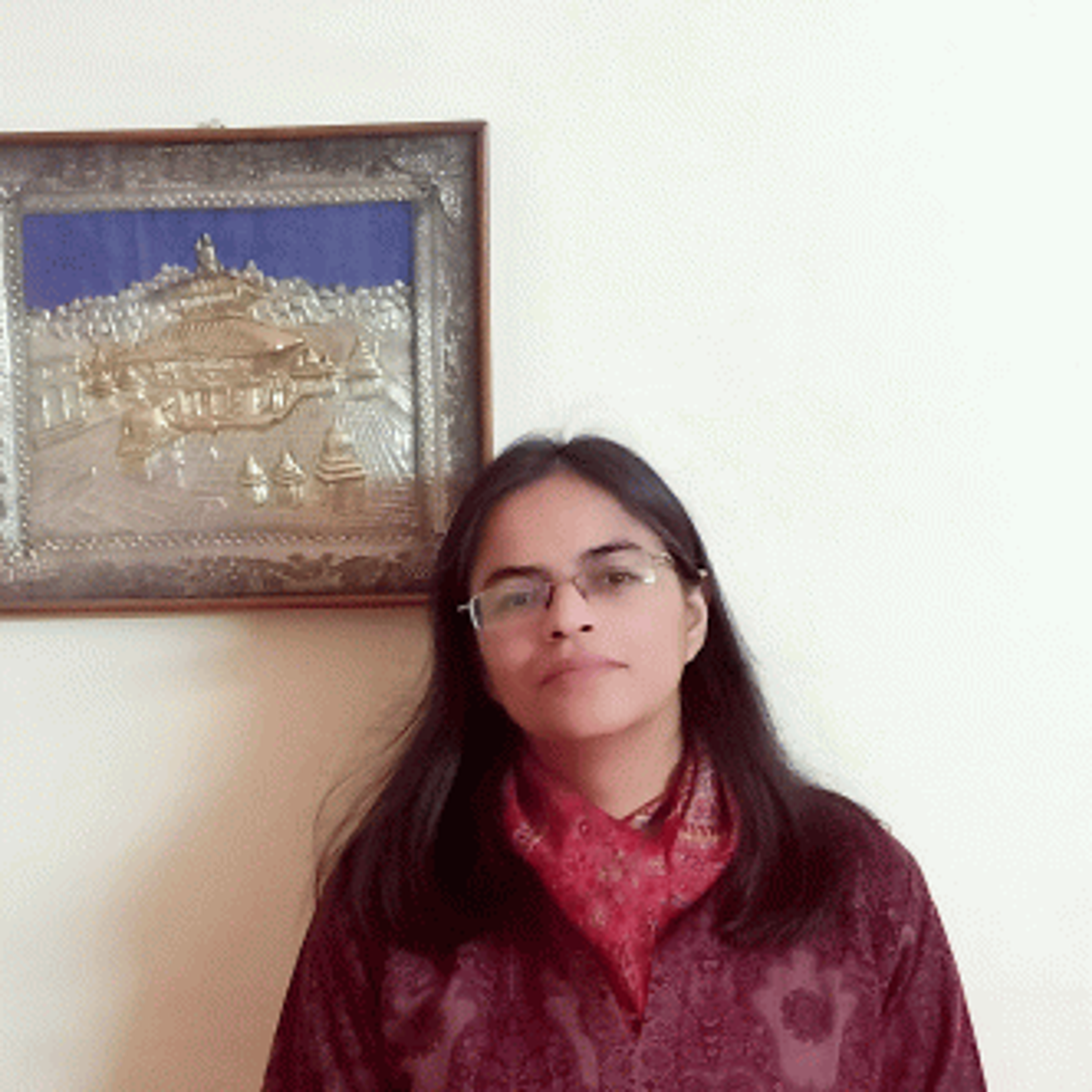 A person with long dark hair and glasses stands indoors, wearing a maroon outfit and a red scarf. A framed artwork is on the wall behind them.