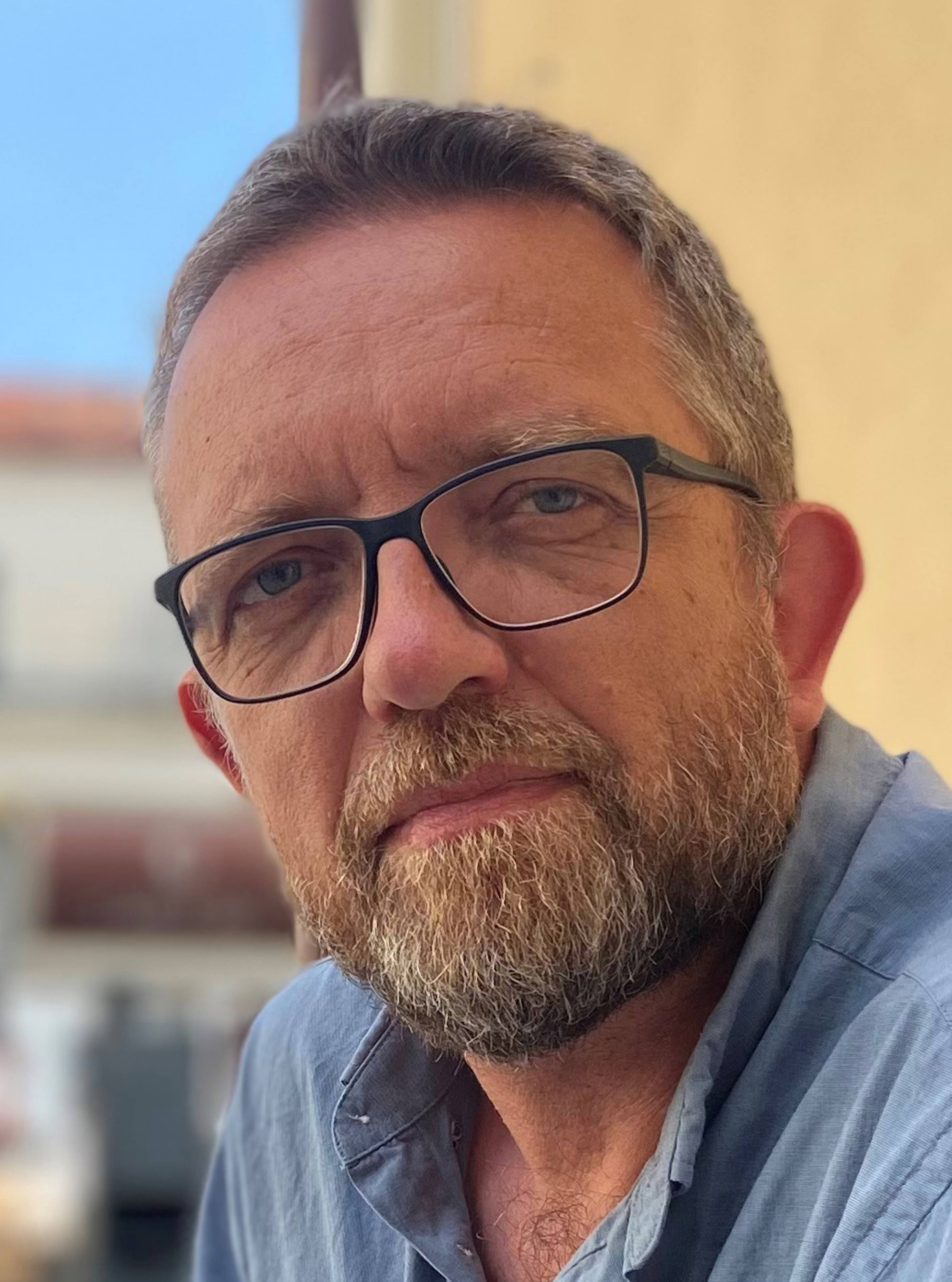 A person with short hair, a beard and glasses looks into the camera. She is wearing a blue shirt. The background is blurred and shows some buildings.