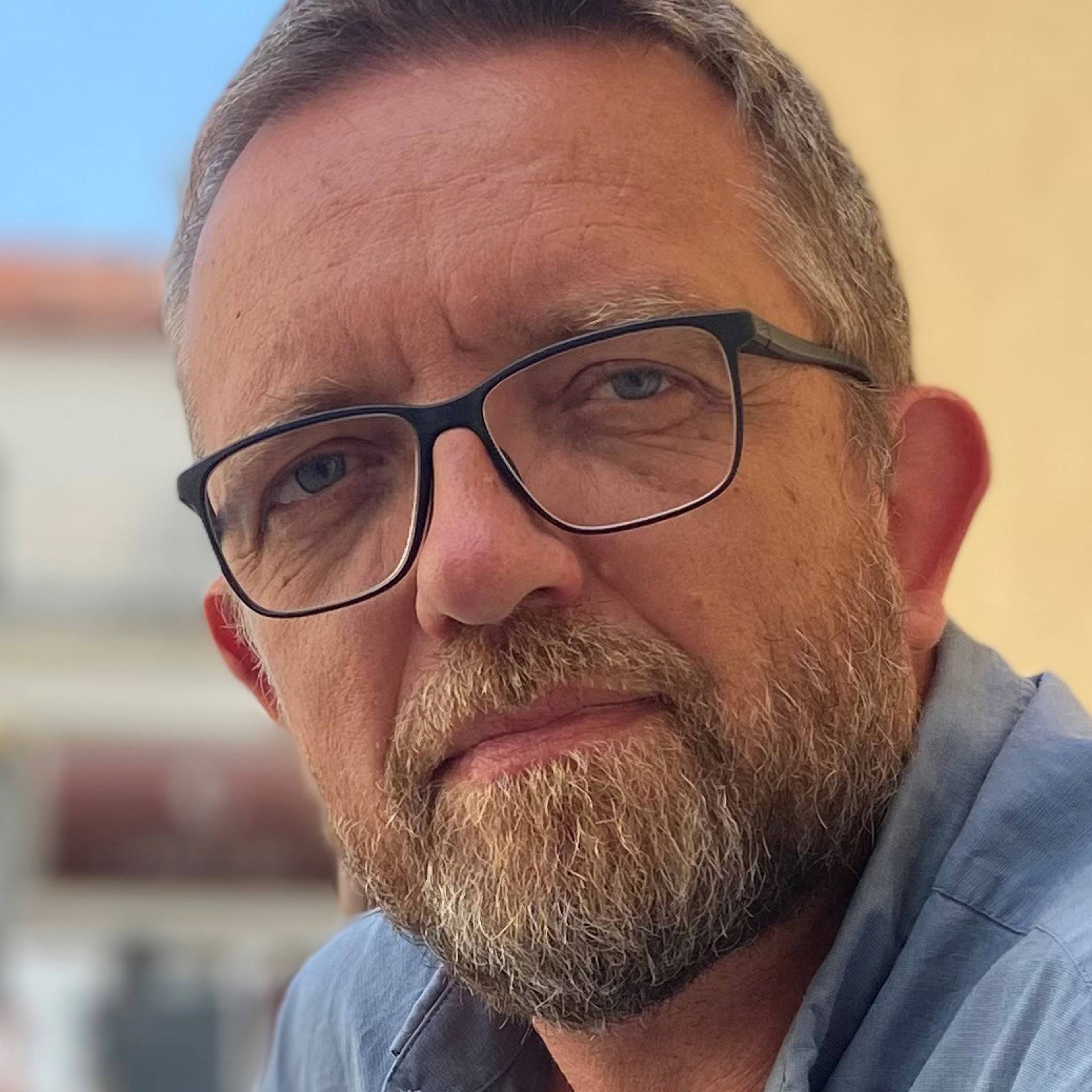 A person with short hair, a beard and glasses looks into the camera. She is wearing a blue shirt. The background is blurred and shows some buildings.