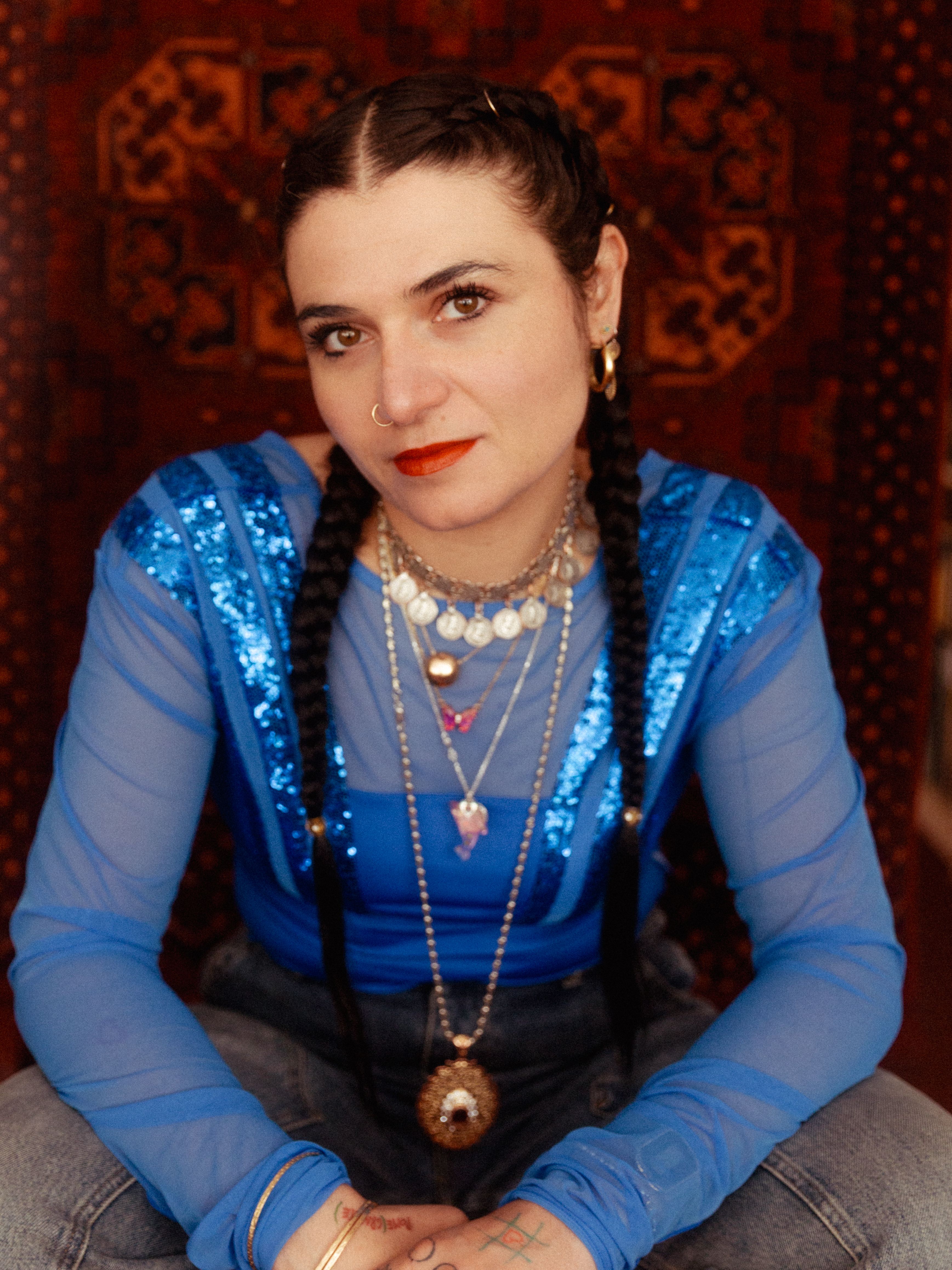 A person with long dark hair styled in braids is seated in front of a patterned background, wearing a sheer blue top with sequins, layered necklaces, and red lipstick.
