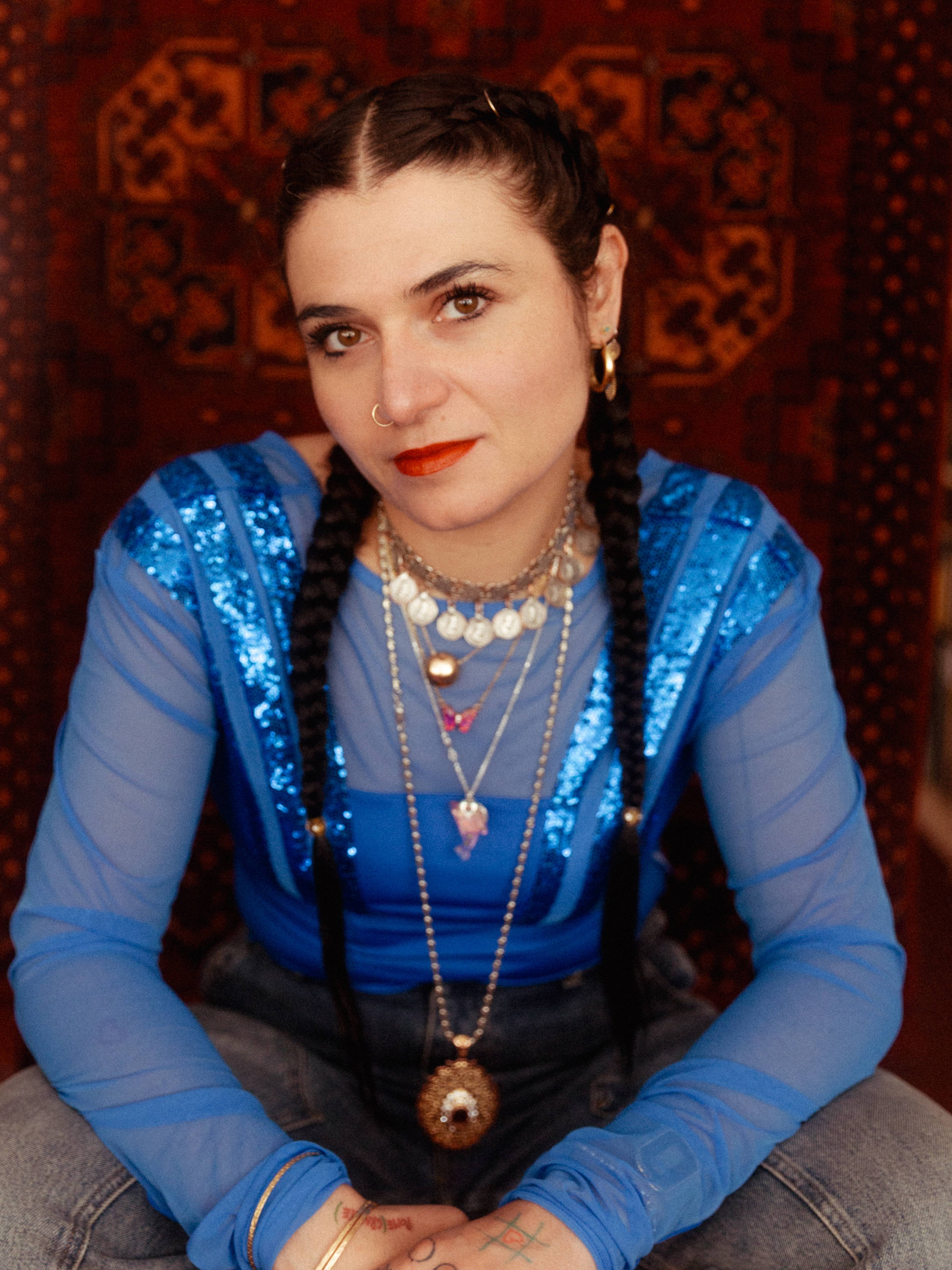A person with long dark hair styled in braids is seated in front of a patterned background, wearing a sheer blue top with sequins, layered necklaces, and red lipstick.